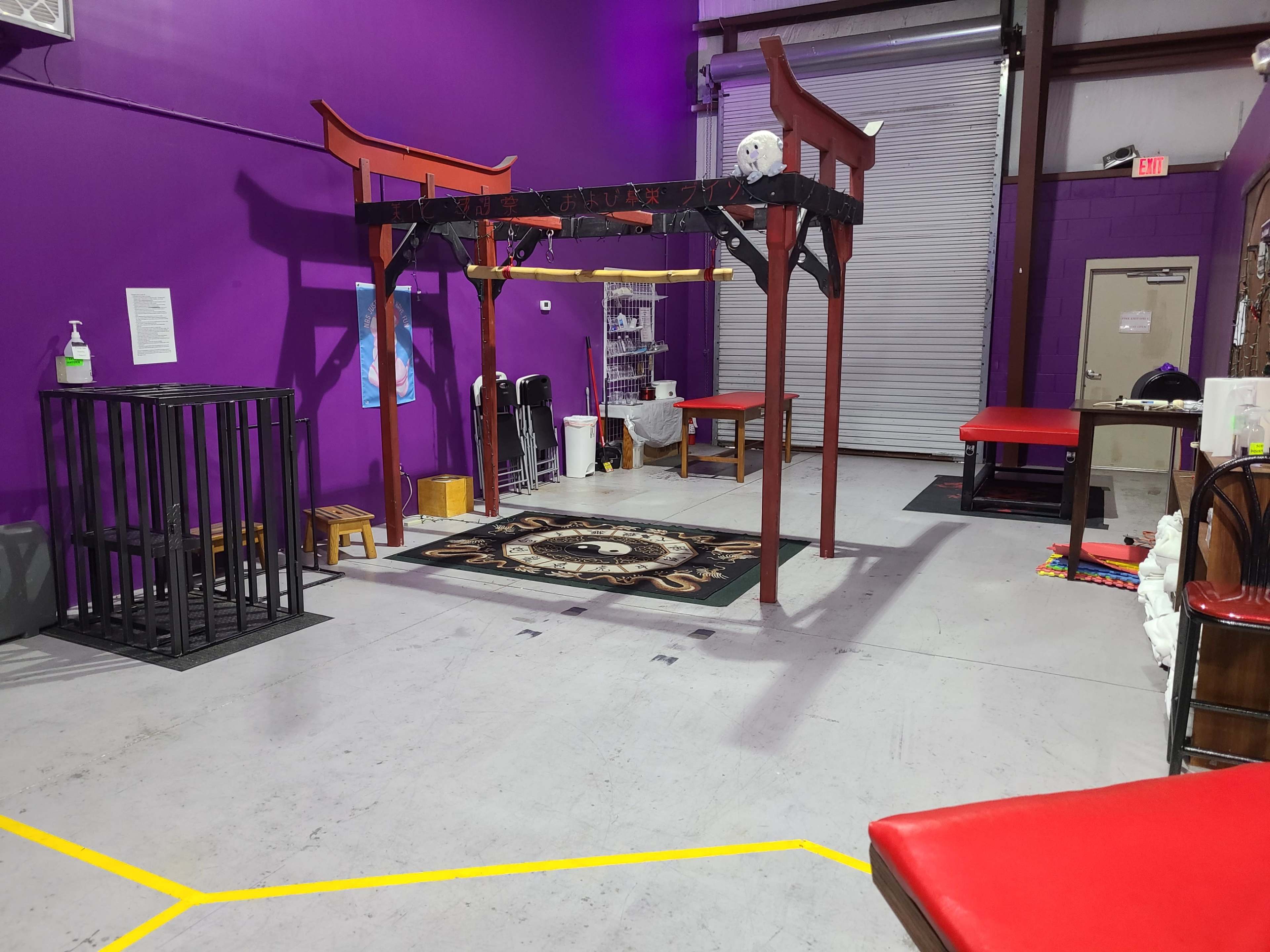 A gym setting with a purple wall, featuring a structure with a wooden frame in the center, a patterned rug on the floor, and various gym equipment and tables along the walls.