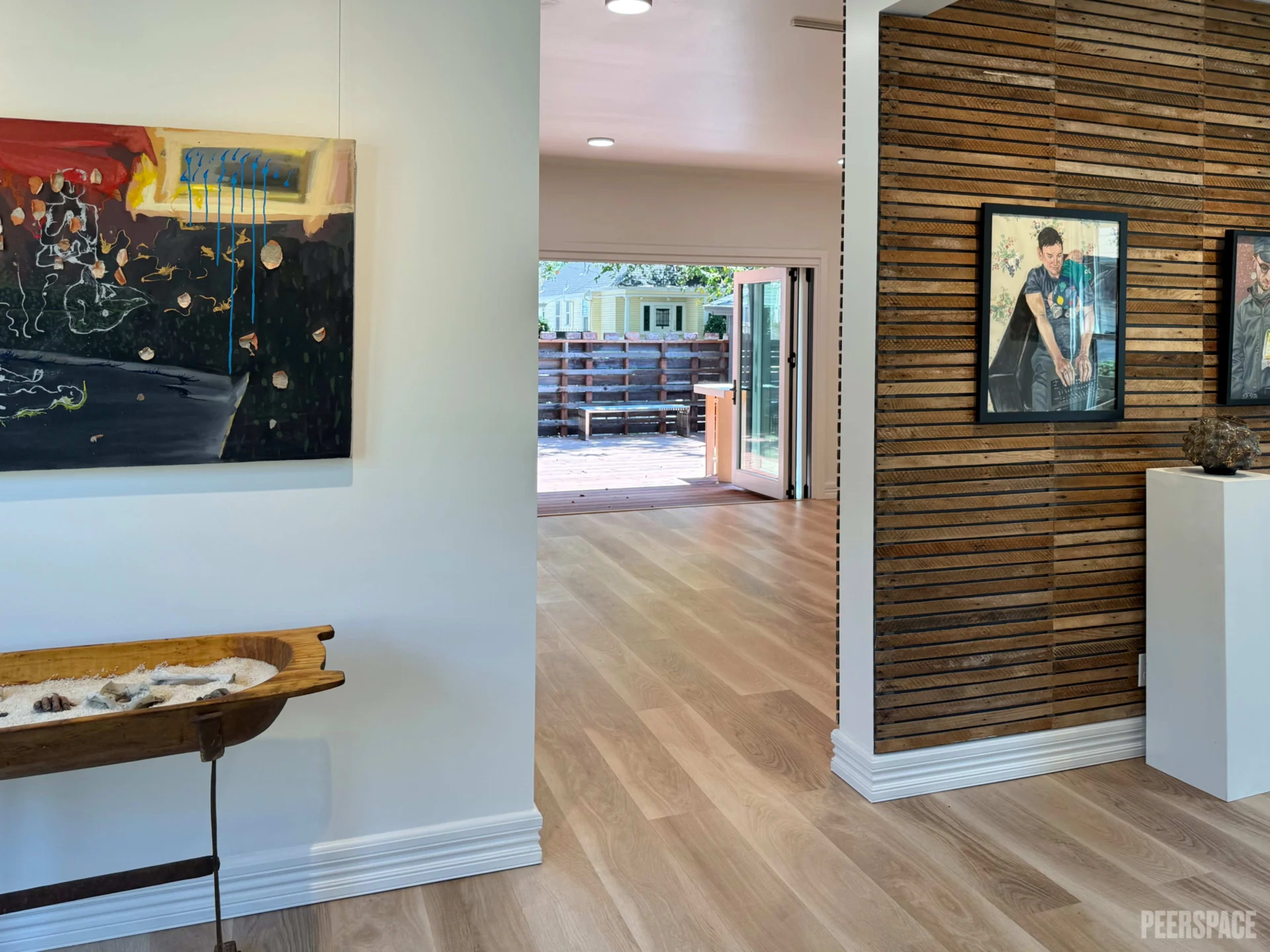 The image shows an interior space featuring a modern art piece on the left and a wooden slatted wall on the right, leading to an open doorway that reveals an outdoor area.
