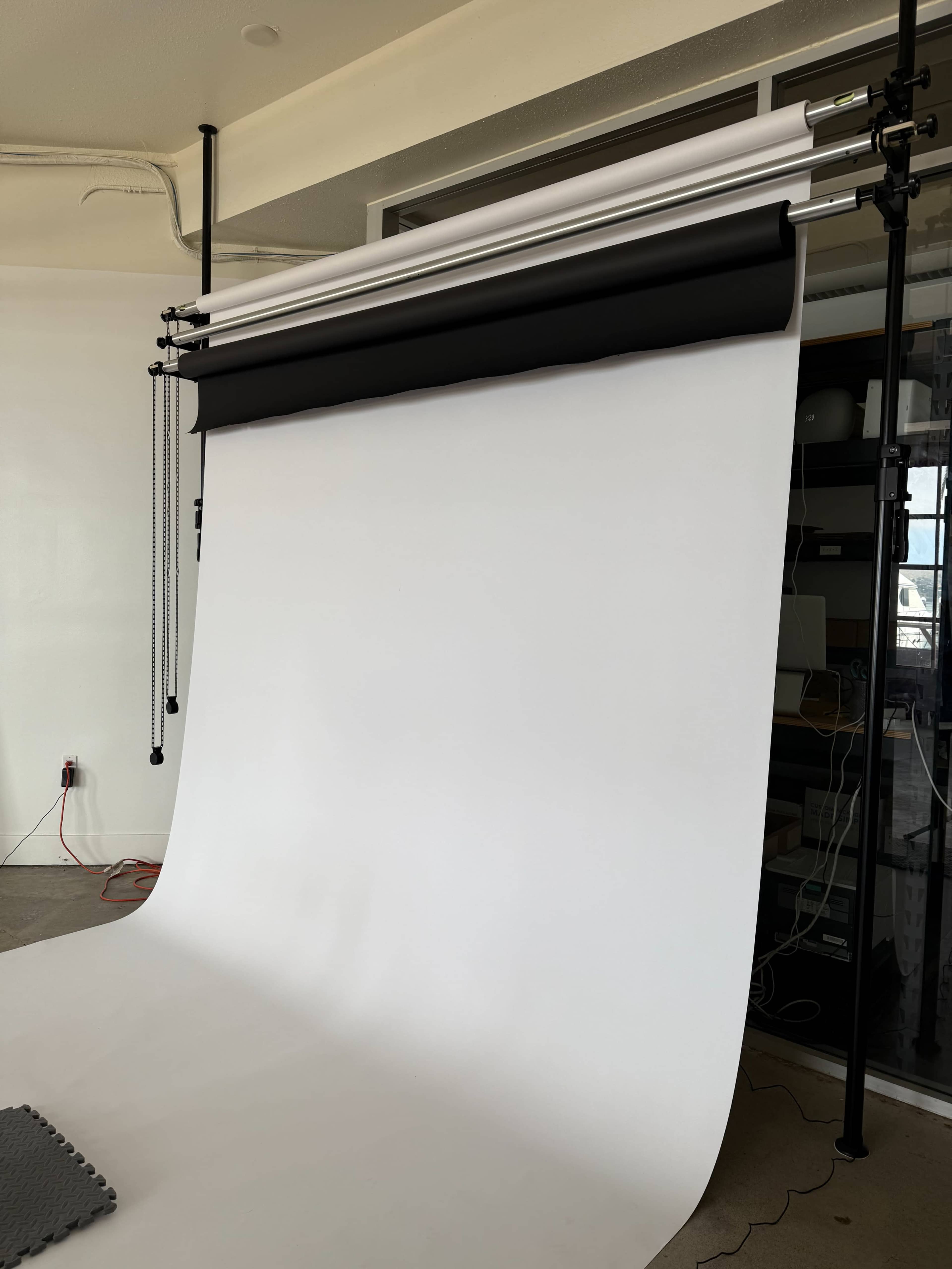 The image shows a photography backdrop setup featuring a white background and rolls of black and other colored materials suspended above it in a studio environment.