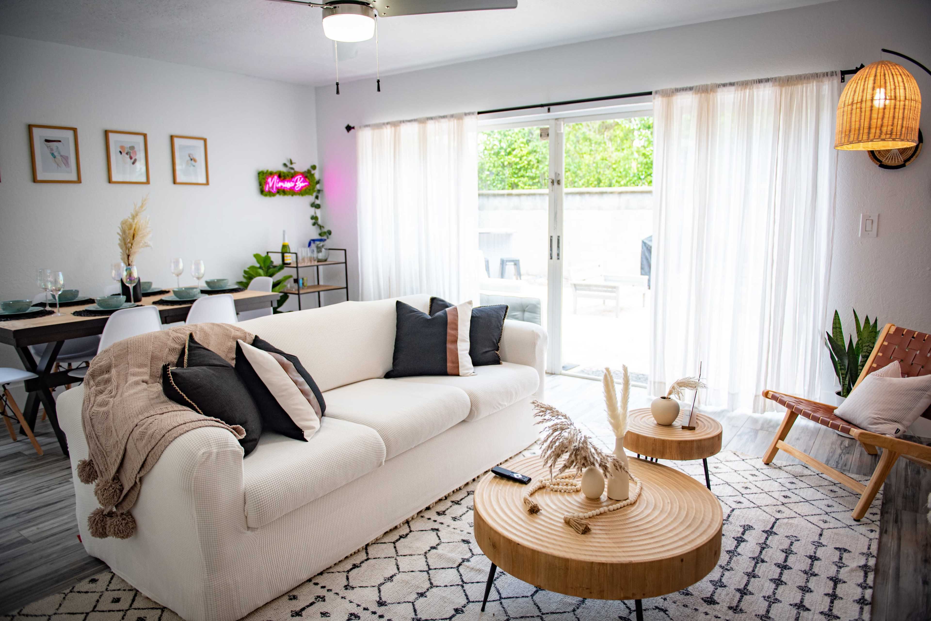 A modern living room featuring a white sofa with black and beige pillows, two wooden coffee tables, and large sliding glass doors that lead to a patio.