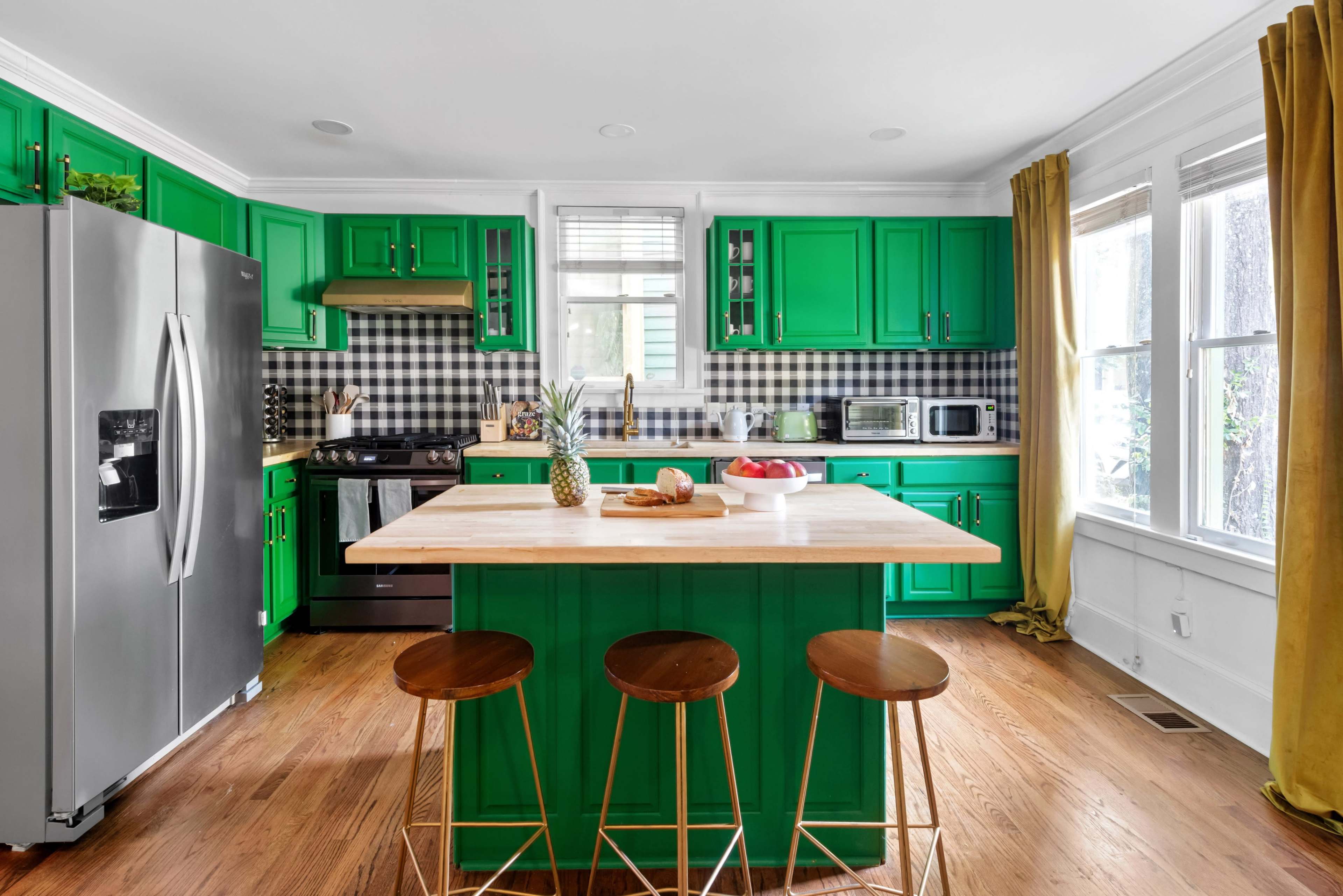 A bright green kitchen features a central wooden island with three stools, surrounded by black and white checkered walls and modern appliances.