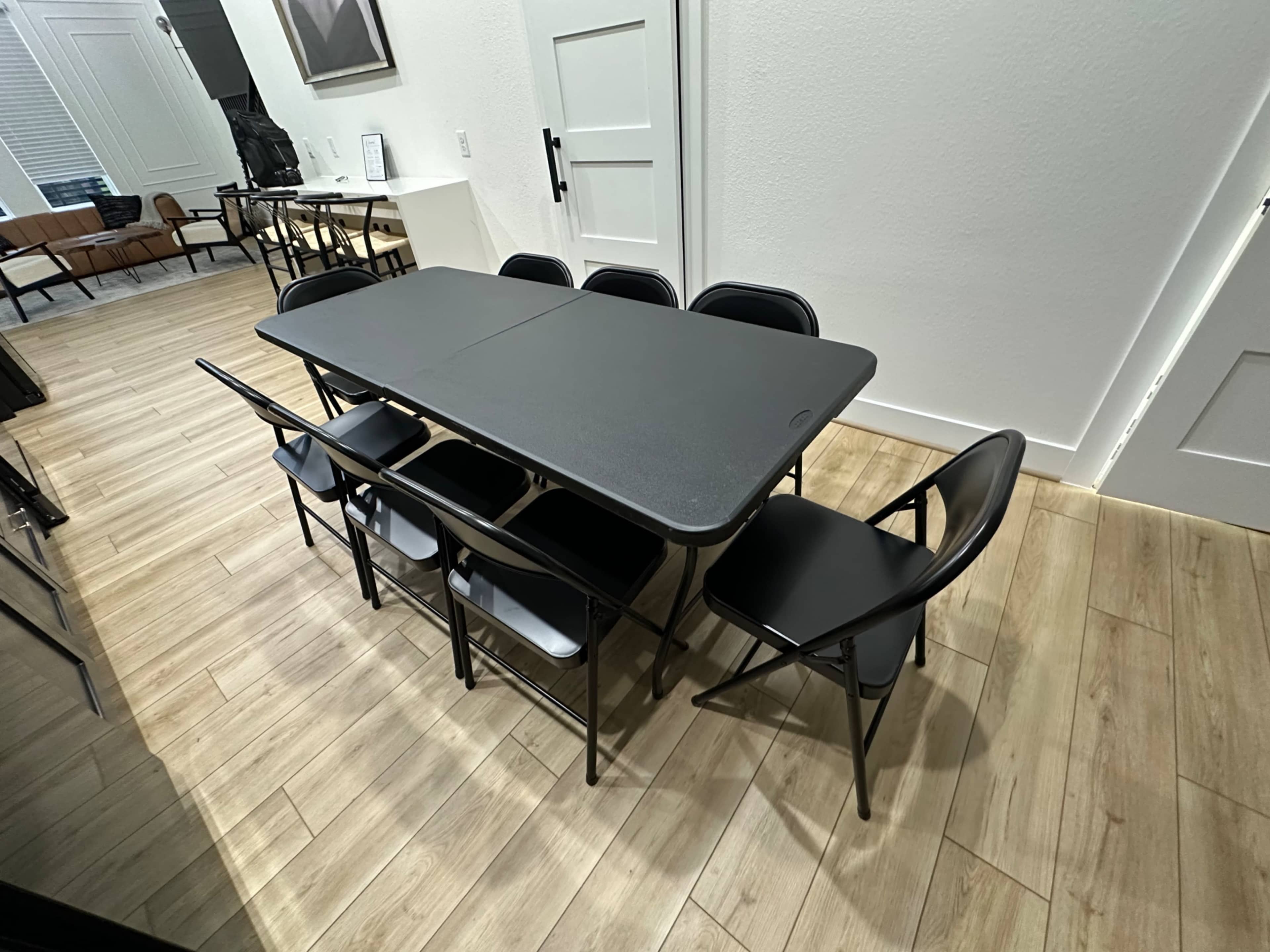 A black folding table surrounded by several black folding chairs is set up in a brightly lit room with wooden flooring.