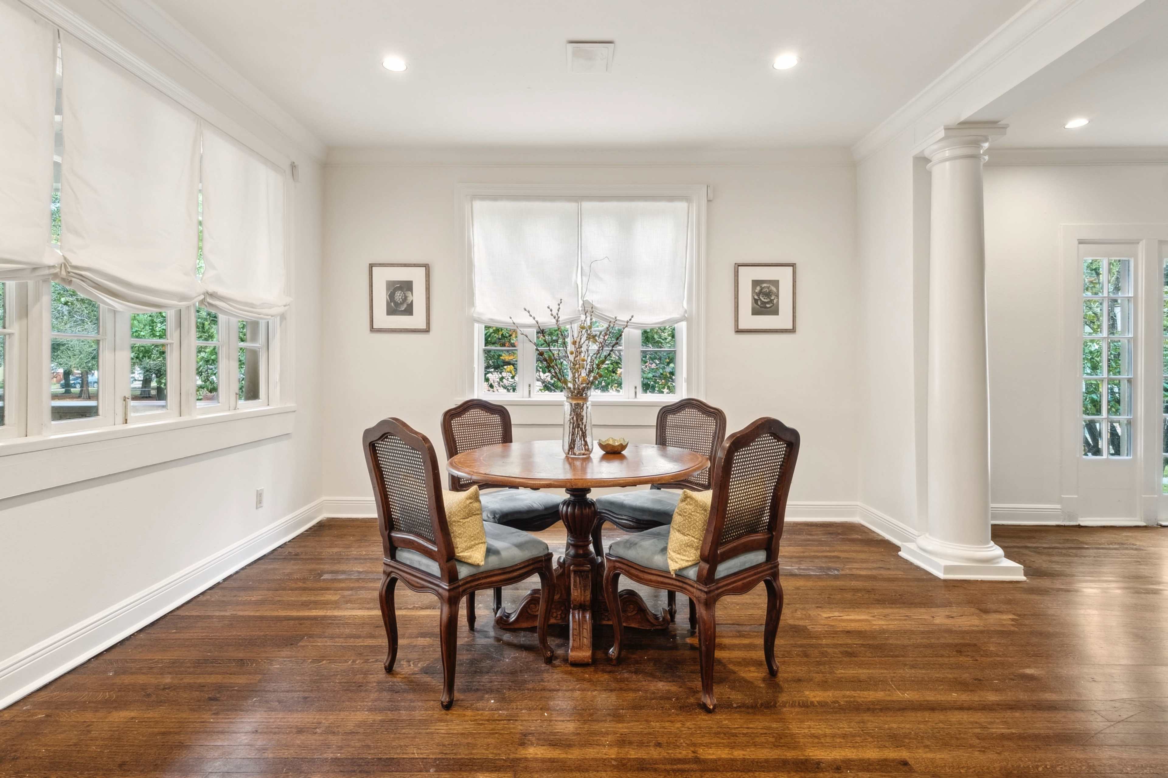 A round wooden dining table with four chairs is set in a well-lit room featuring white walls and large windows adorned with light-colored shades.