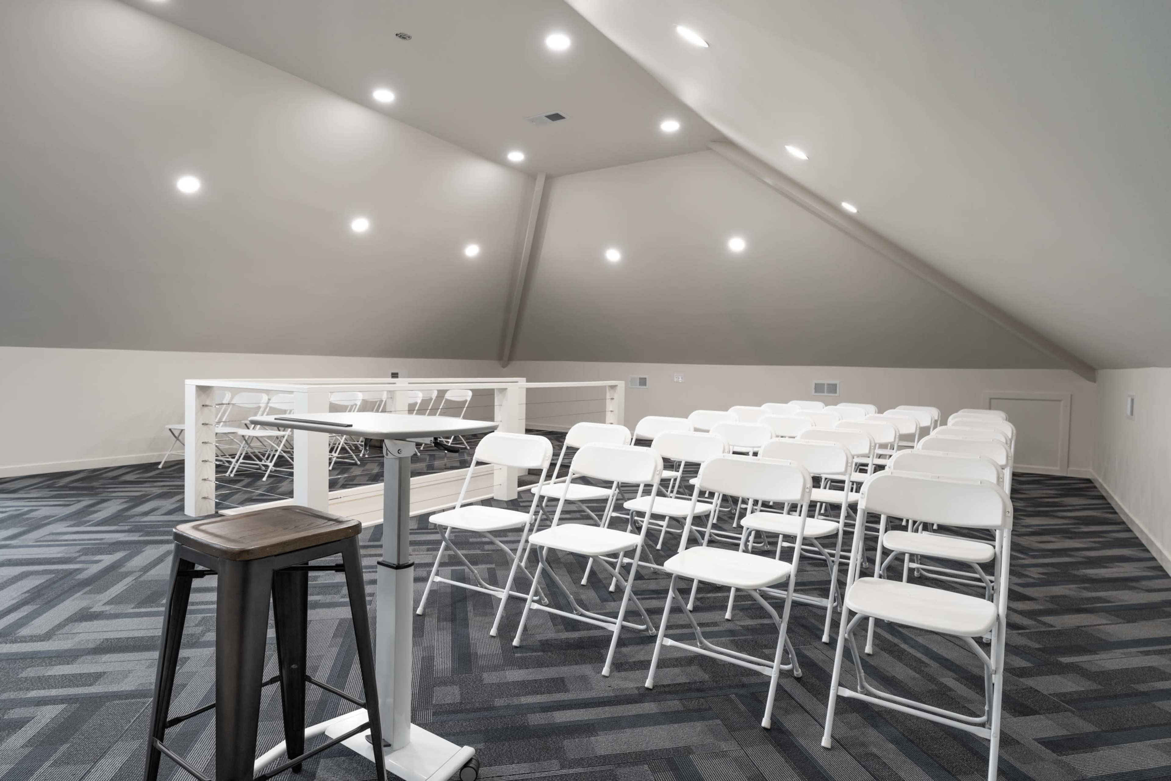 A spacious attic room features rows of white folding chairs set up for an event, with a small podium in the foreground.