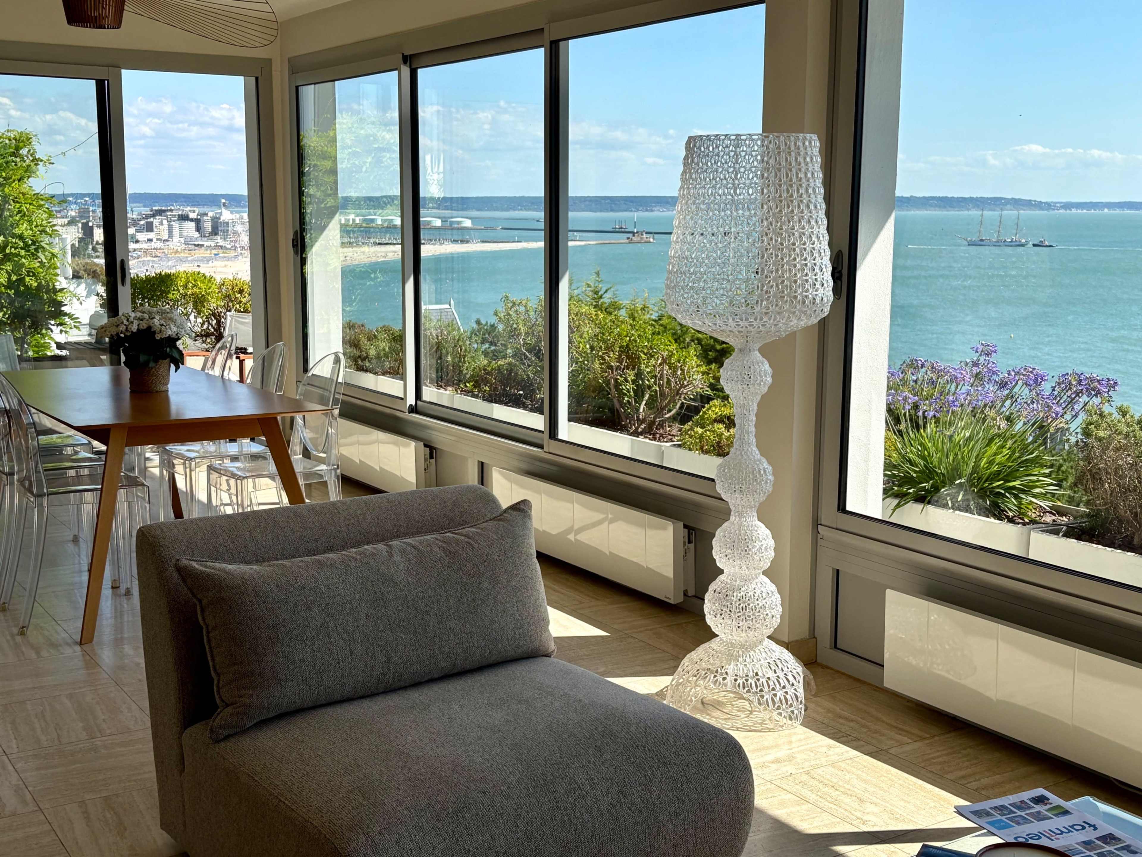 A bright room features a transparent lamp, a modern sofa, and large windows showcasing a coastal view with boats in the distance.