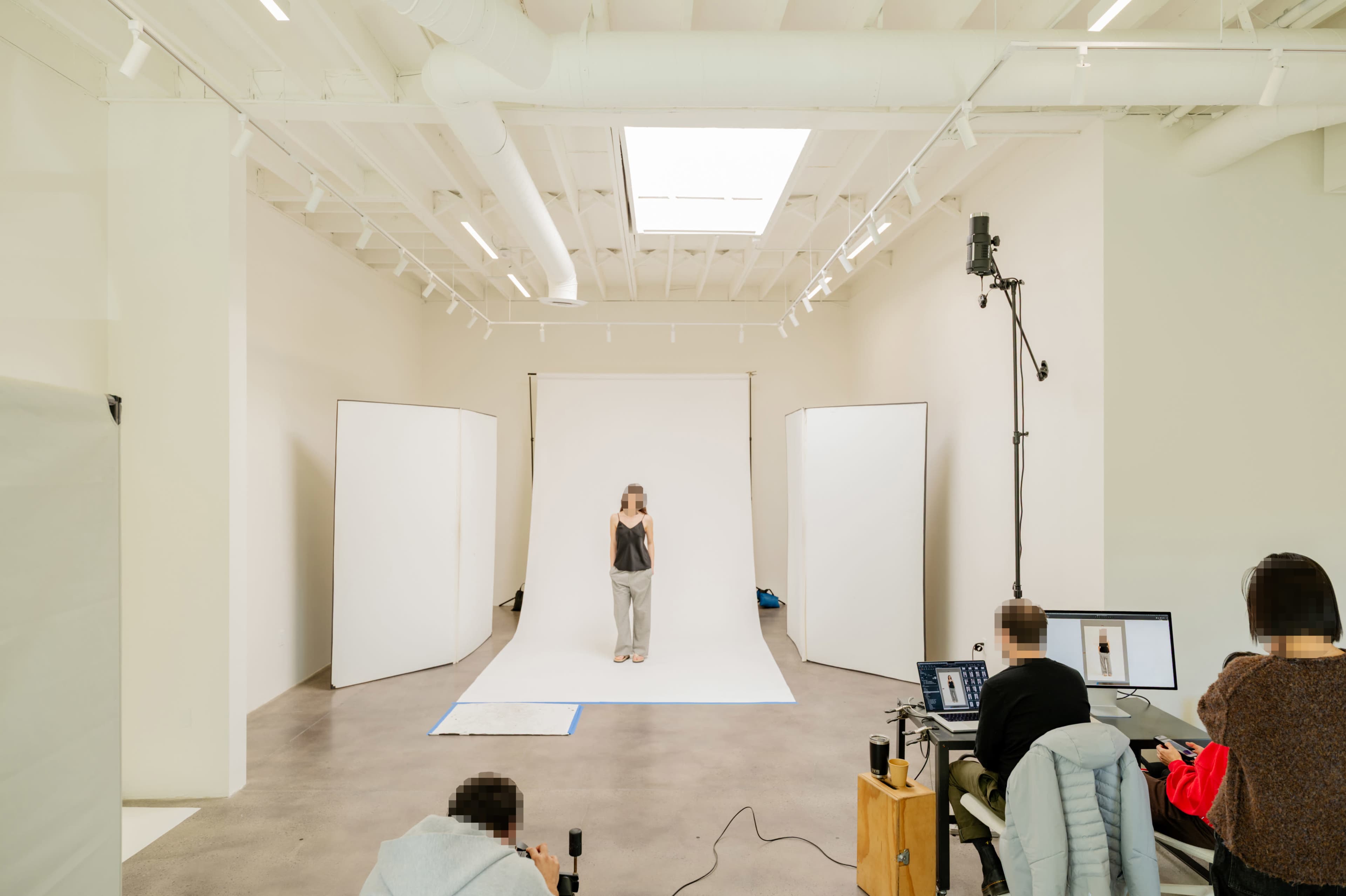 A photography studio features a model standing in front of a plain backdrop, with photographers and equipment set up nearby.