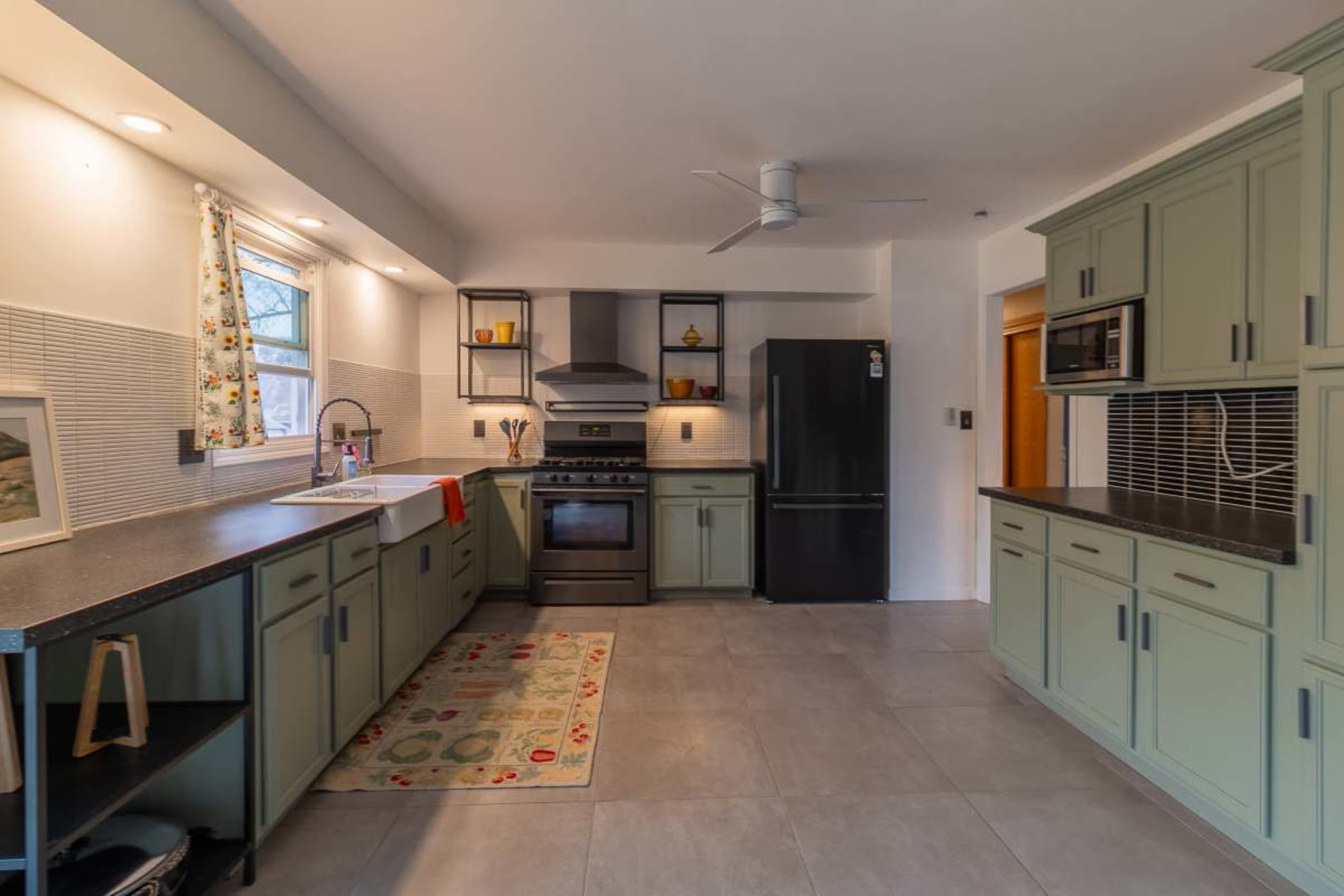 The image shows a modern kitchen with green cabinets, stainless steel appliances, and a countertop with a colorful rug on the tiled floor.