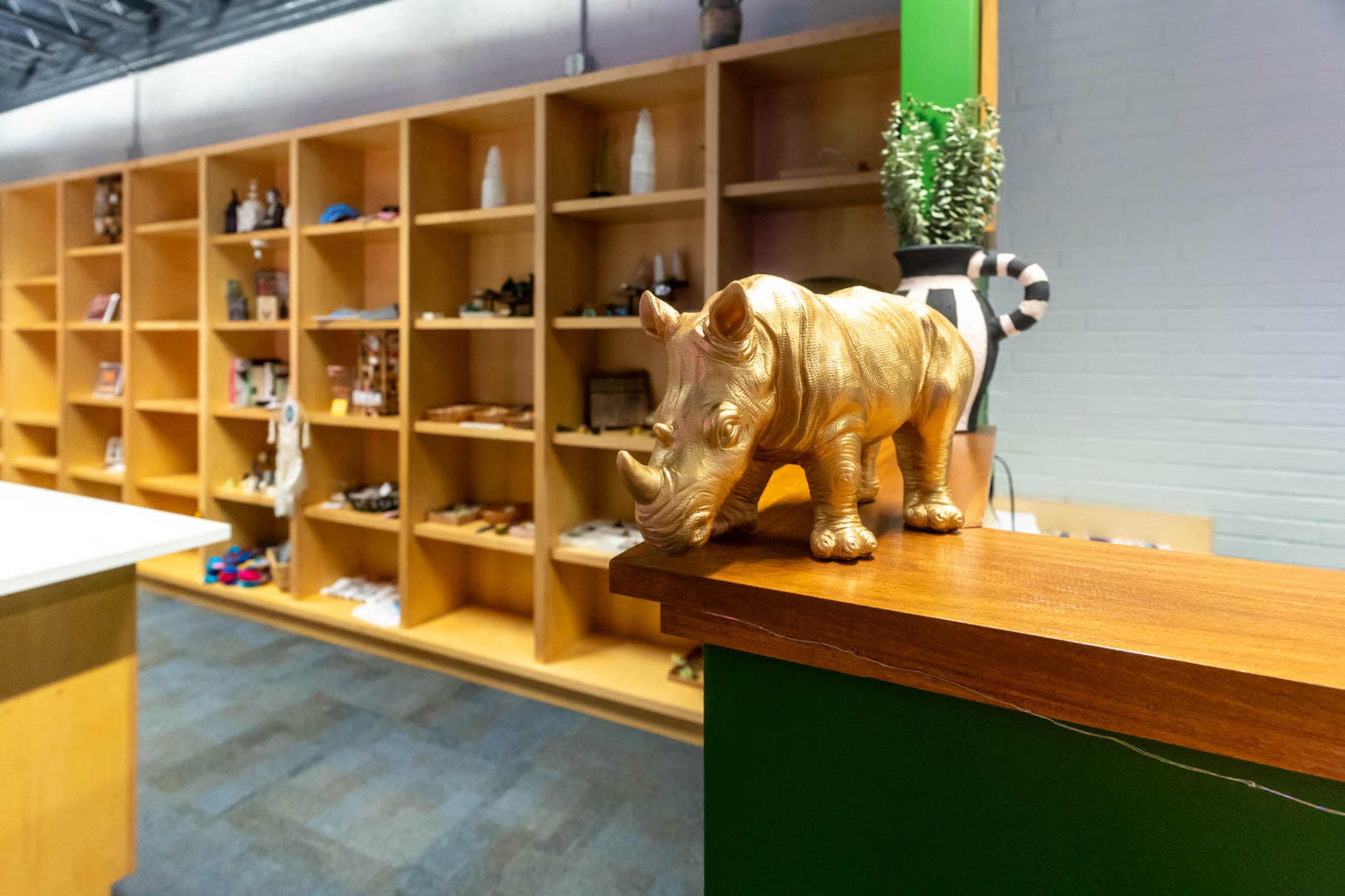 A golden rhinoceros sculpture sits on a wooden ledge in front of a shelf displaying various items.