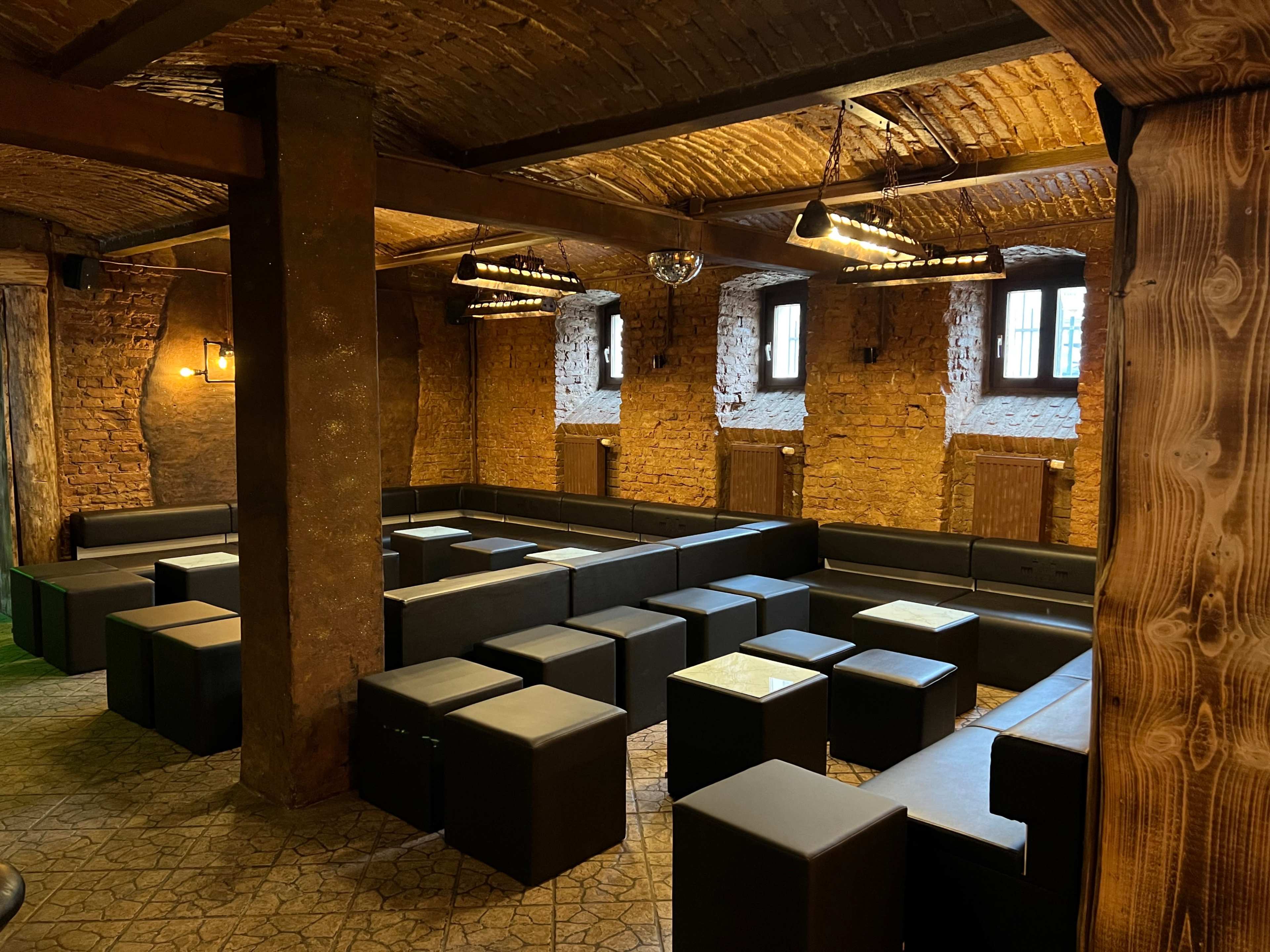 A dimly lit lounge area with black seating cubes arranged around small glass-topped tables against a rustic brick wall.