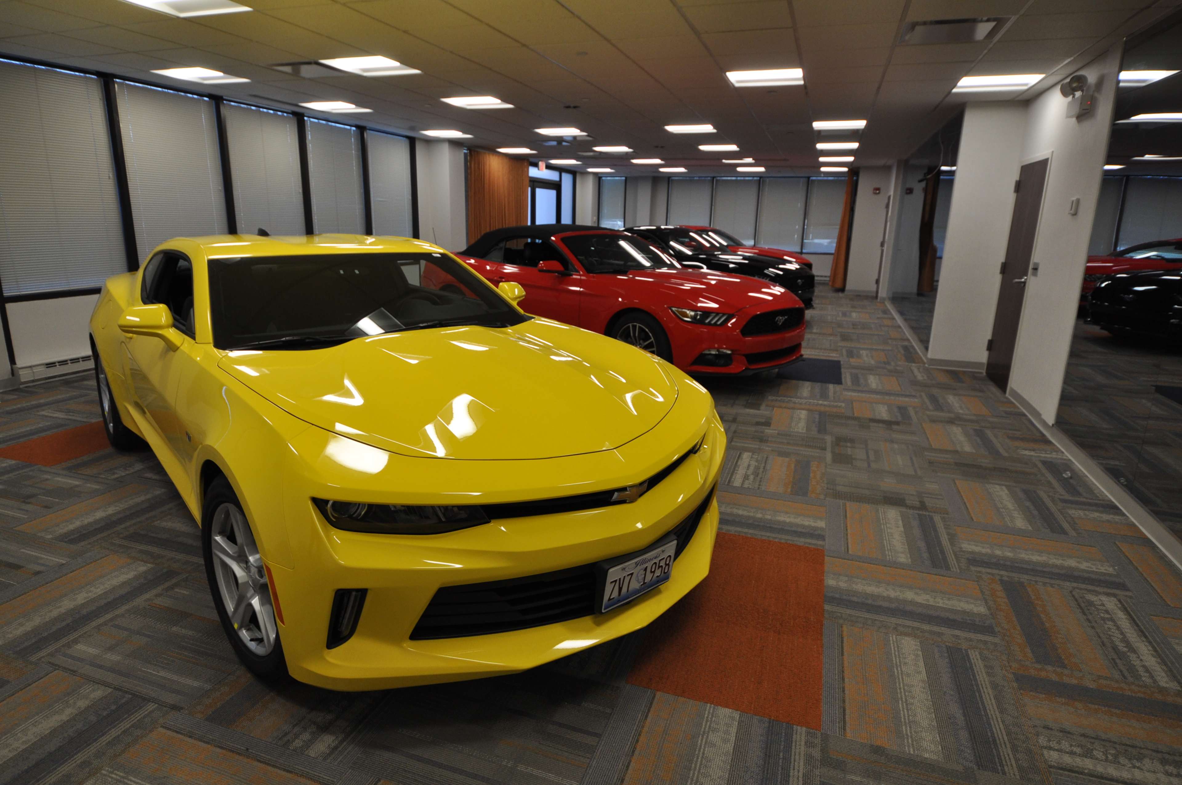 A yellow car is parked in the foreground of an office space, with a red car and a black car visible in the background.