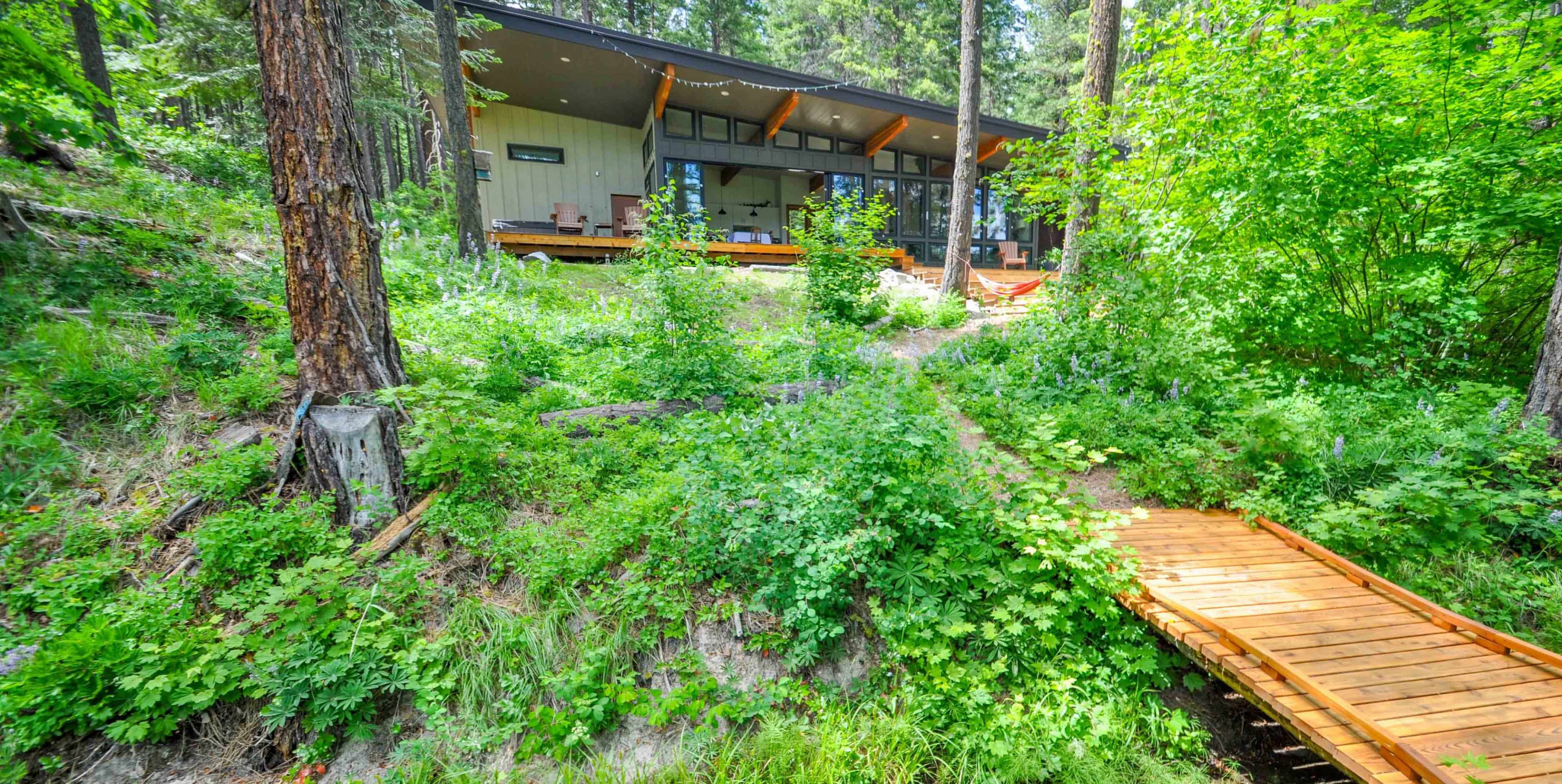 A wooden walkway leads to a modern house nestled among lush greenery and trees.