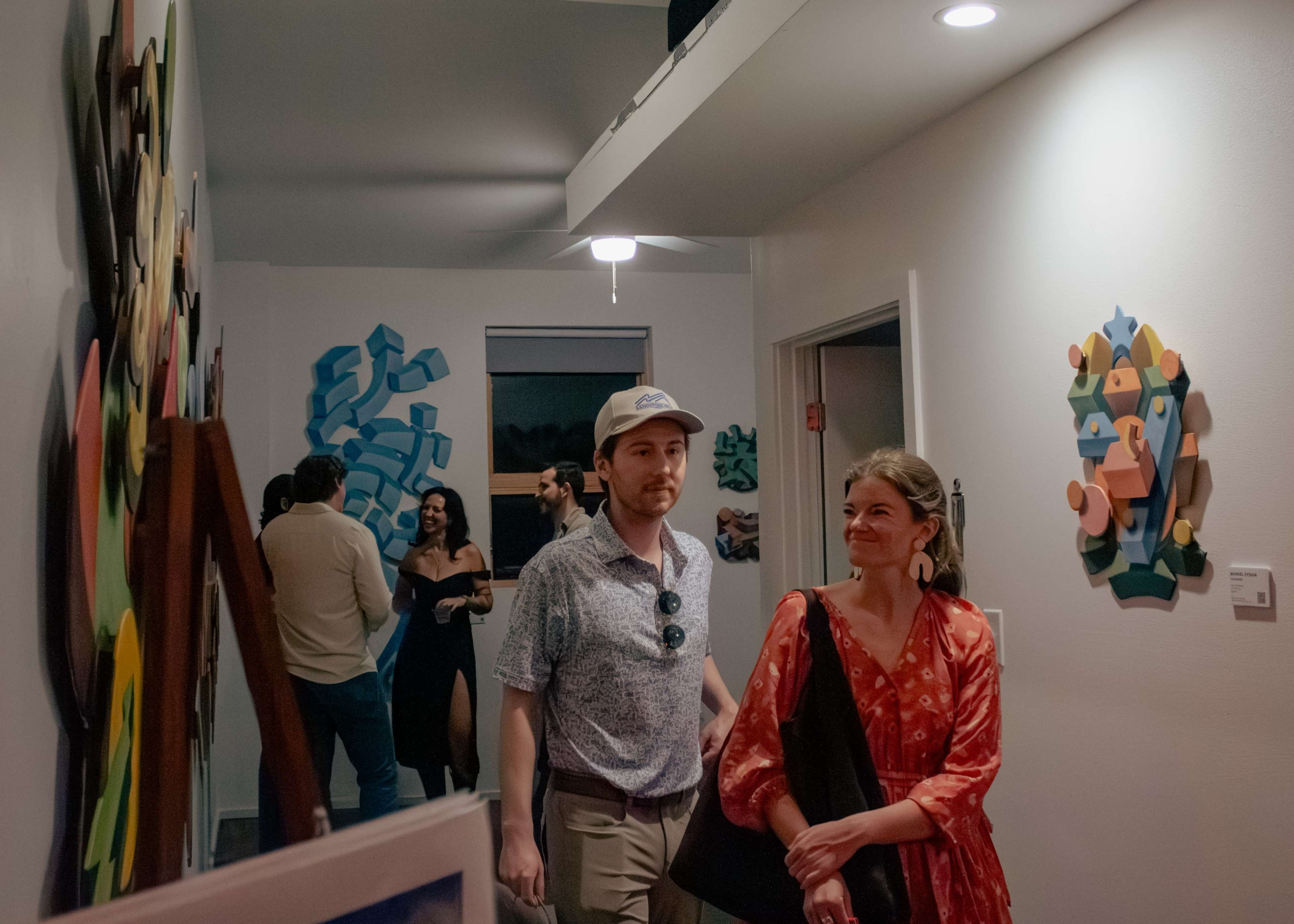 A man and a woman walk down a hallway adorned with colorful artwork, while other people are visible in the background.