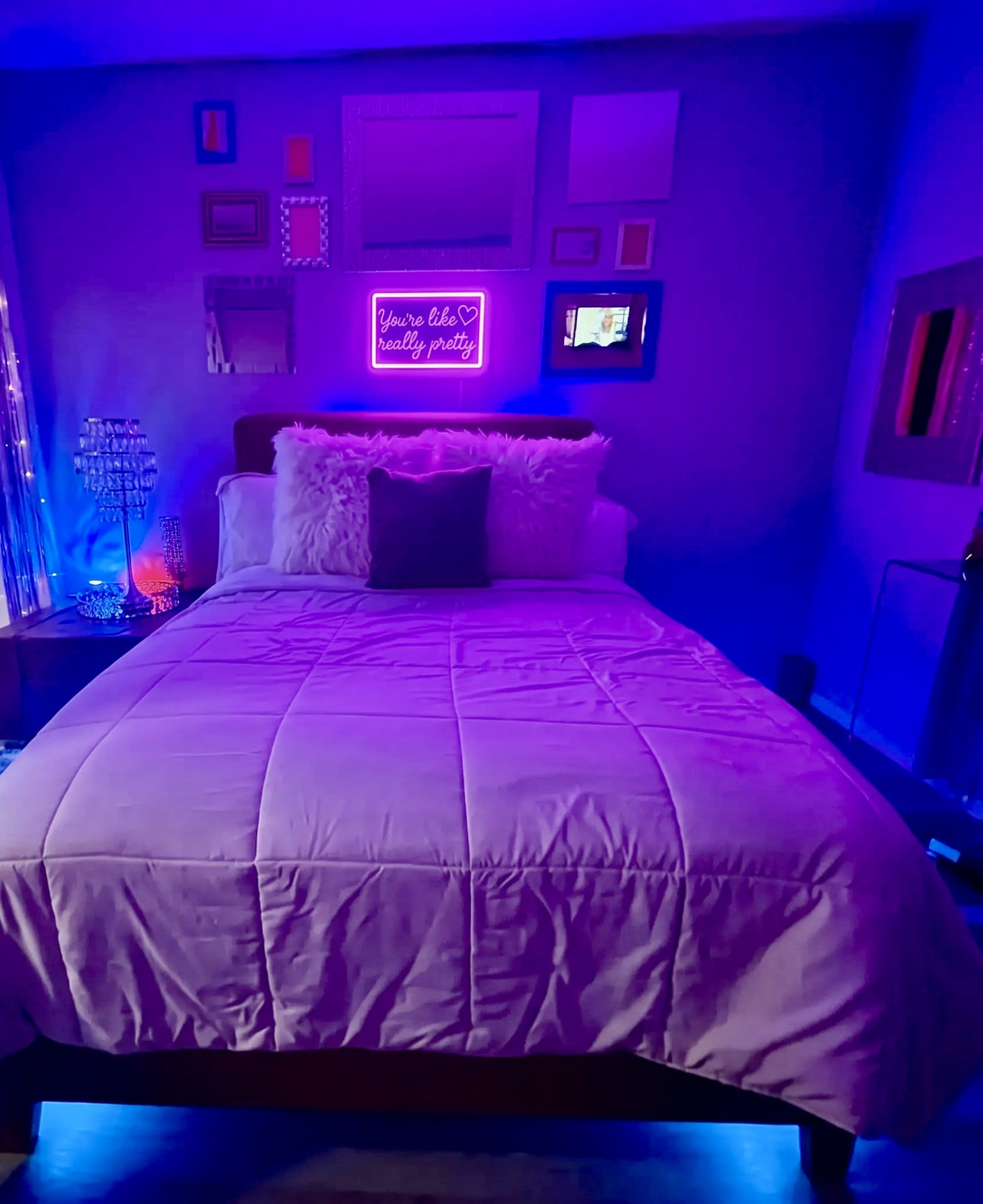 A neatly made bed with a white comforter and decorative pillows is positioned in a dimly lit room adorned with framed pictures and purple lighting.