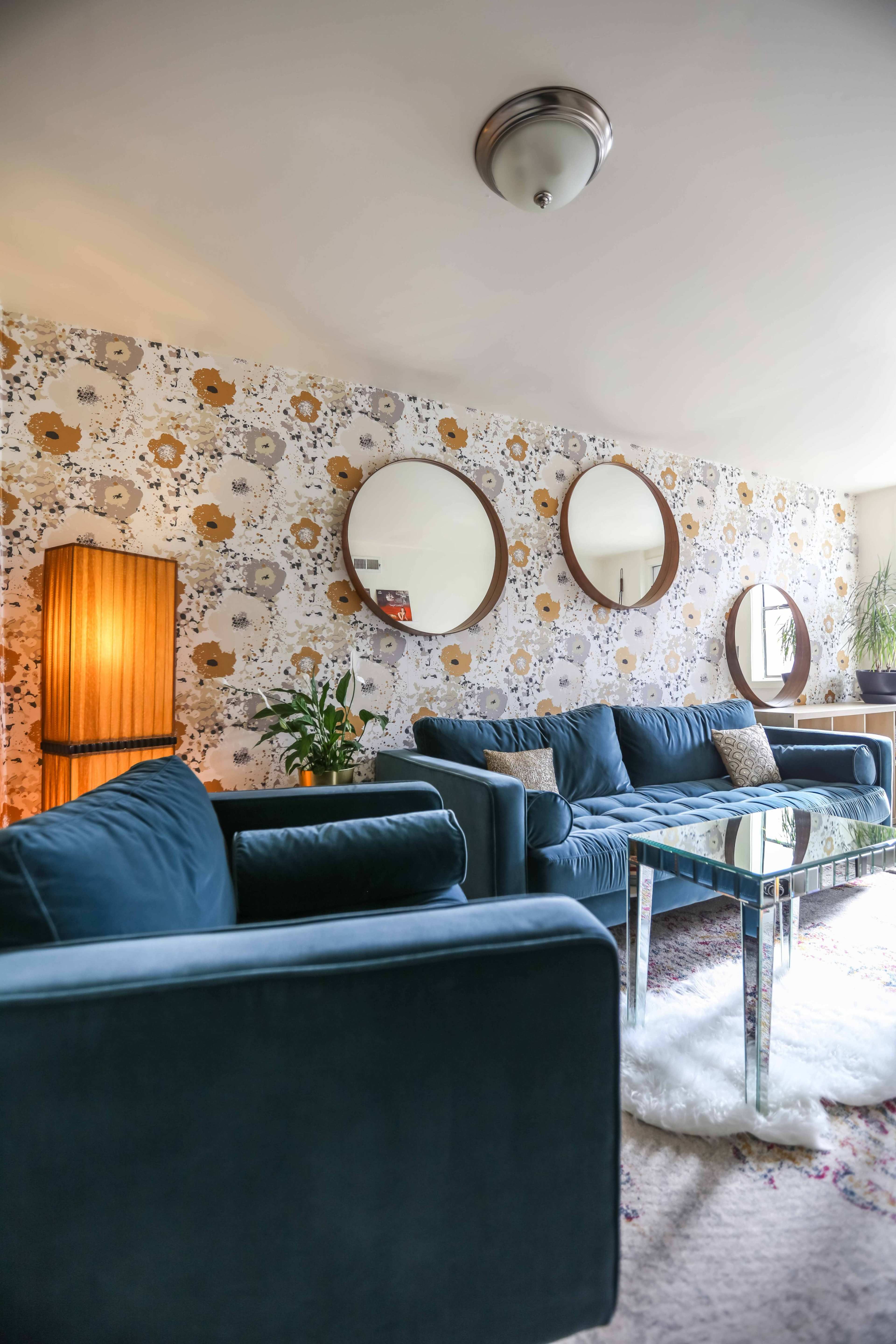 A cozy living room features a floral wallpaper, a pair of round mirrors, a glass coffee table, and blue sofas.