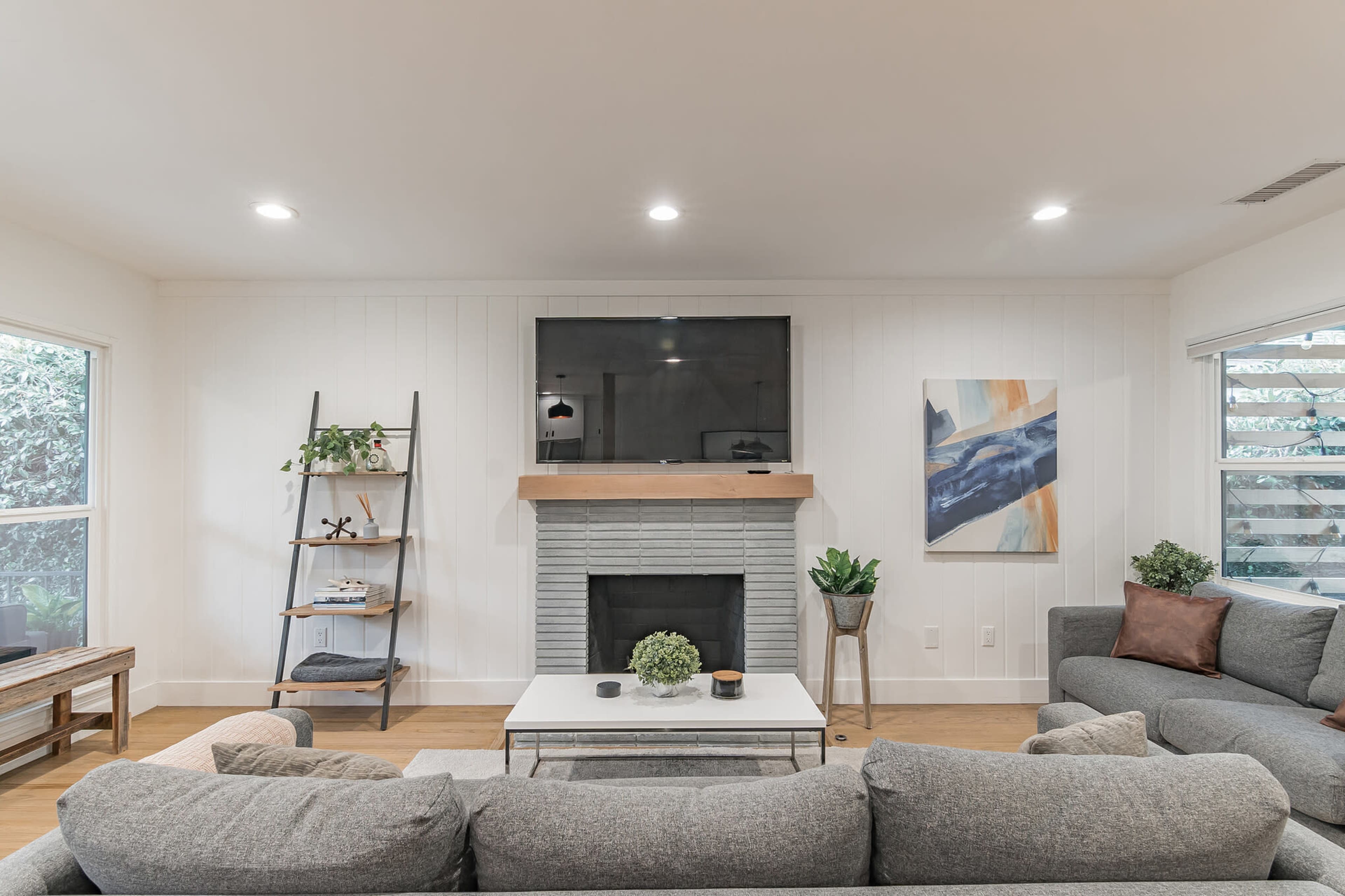 A modern living room features a gray sectional sofa, a white coffee table, a stone fireplace with a mounted TV above it, and a wooden shelf displaying plants and decorative items.