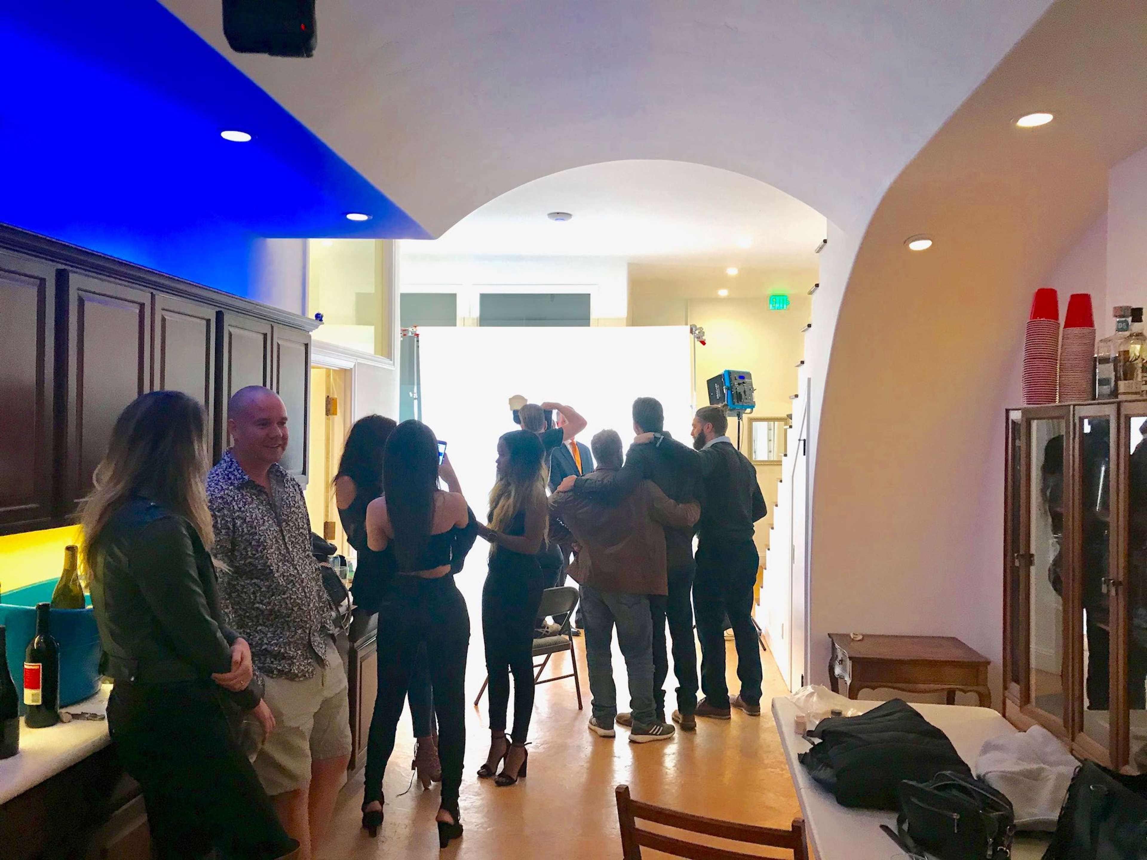 A group of people stands in a kitchen, while another group poses for a photo in front of a bright backdrop.