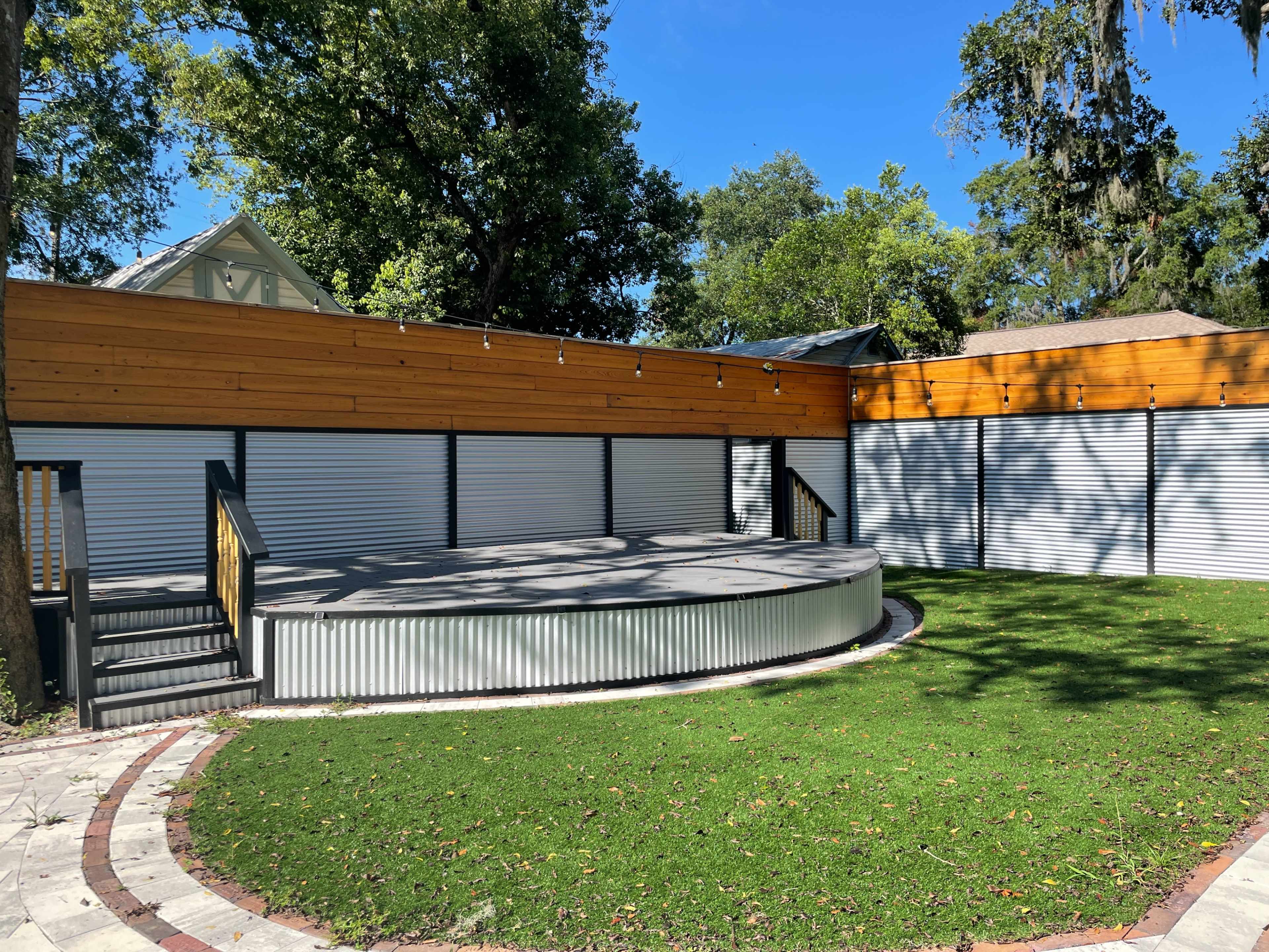 The image shows an outdoor space featuring a circular stage surrounded by wooden walls and a well-maintained grassy area.