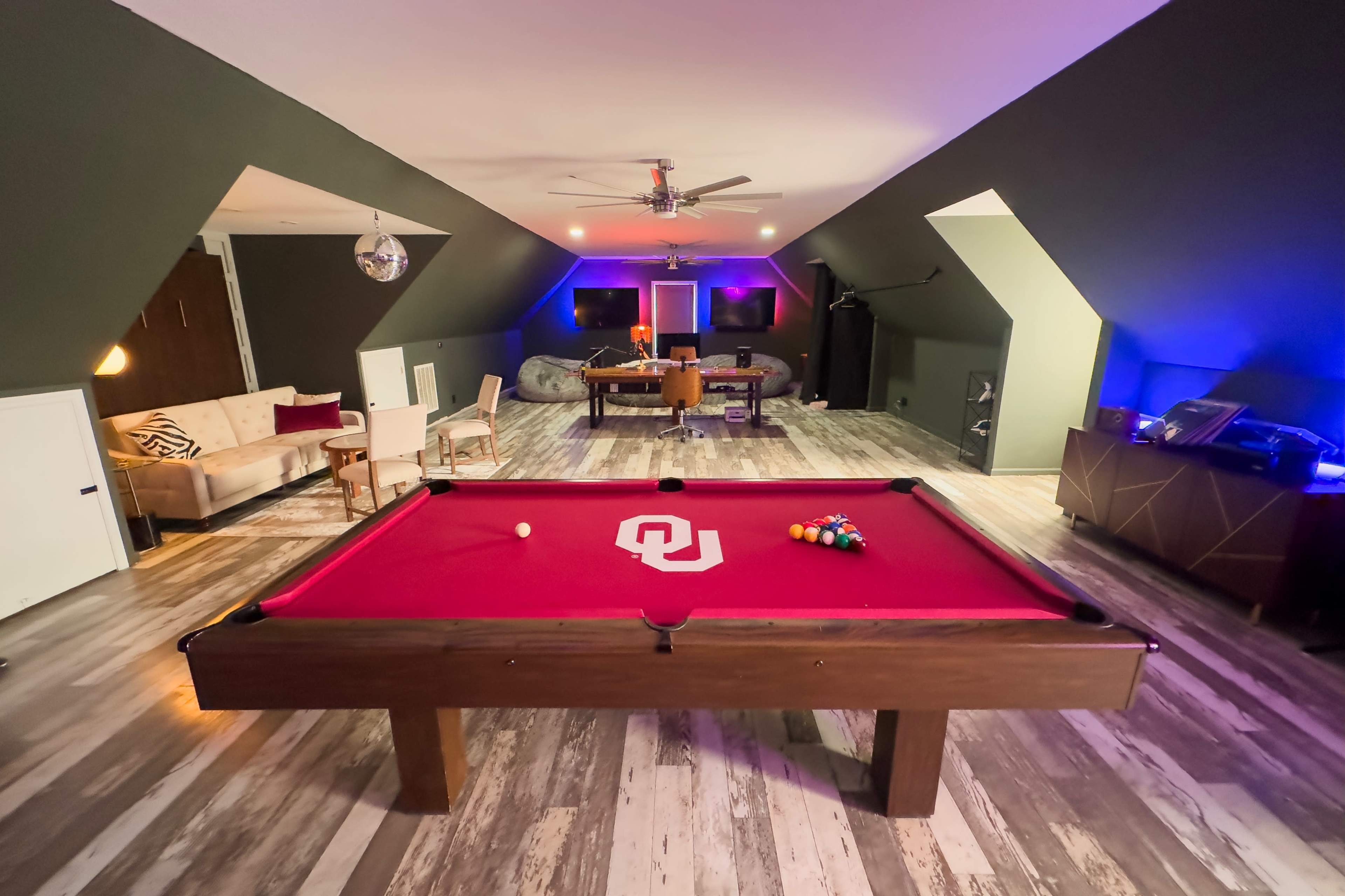 The image shows a game room with a red pool table at the center, surrounded by modern seating and entertainment areas under dim lighting.