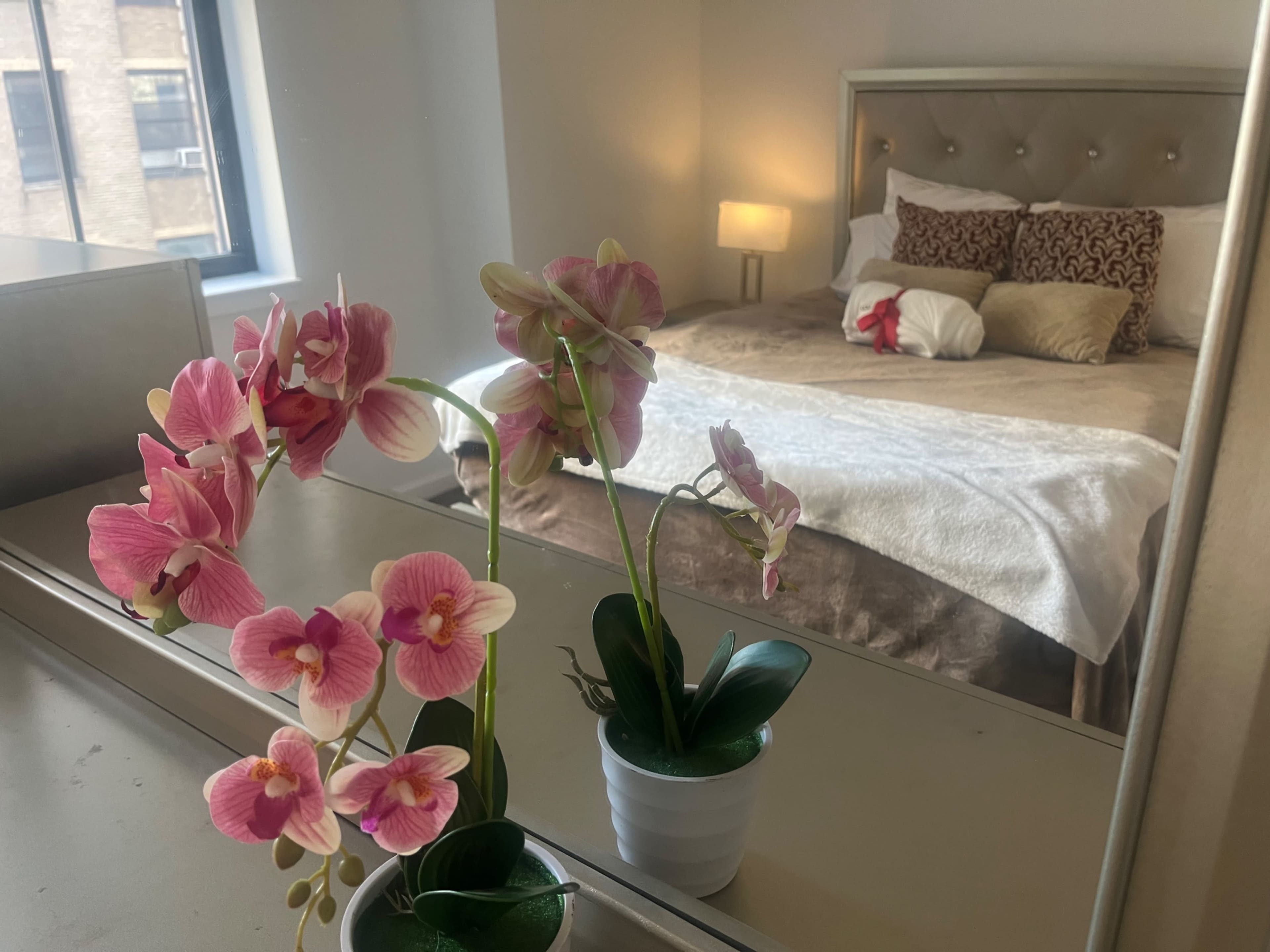 A mirror reflects a bedroom with a bed adorned in neutral linens, while pink orchids sit in a pot on a dresser.
