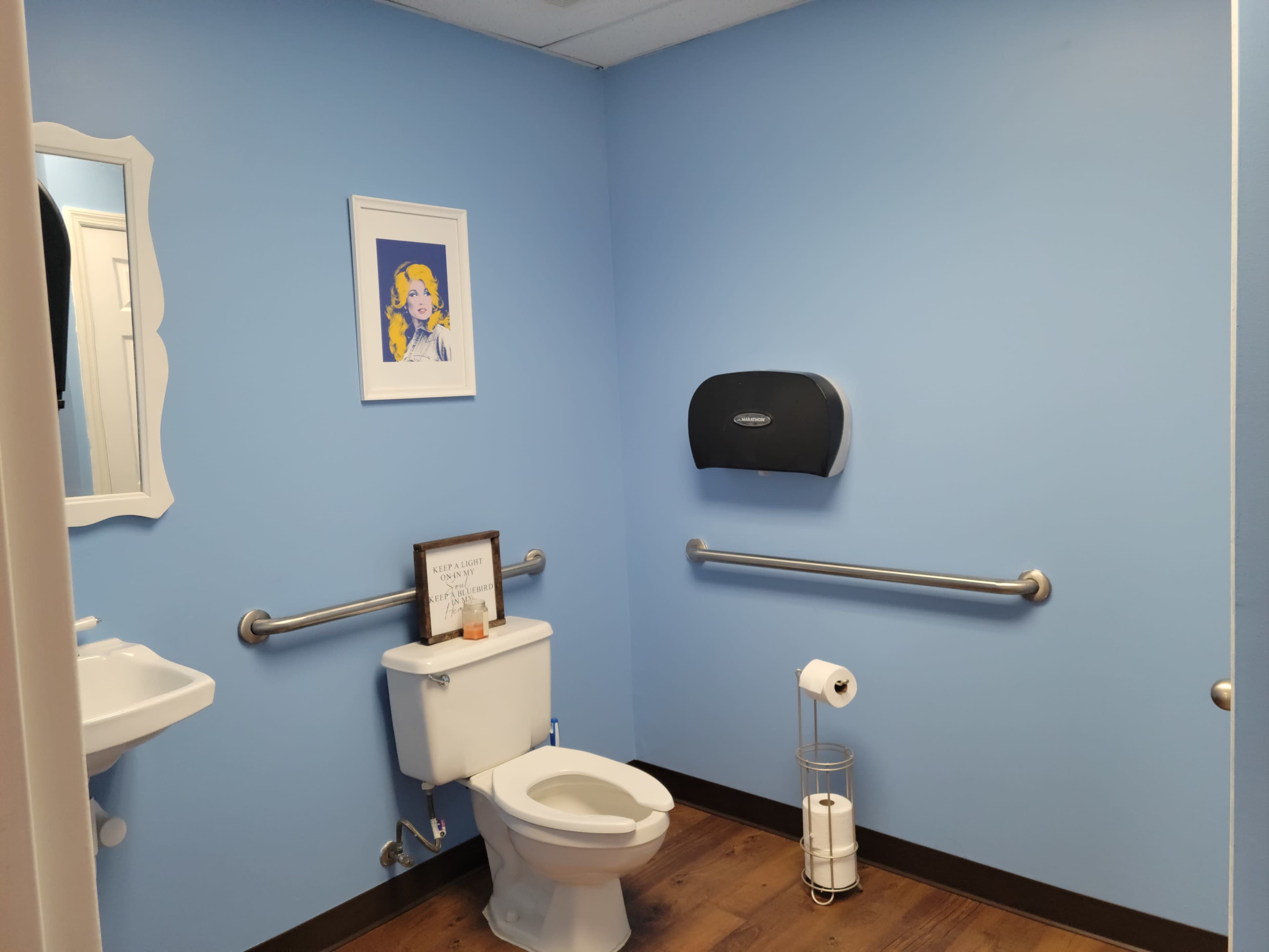 A restroom with blue walls, a toilet, a sink, and some decorative elements including a framed picture and a paper towel dispenser.