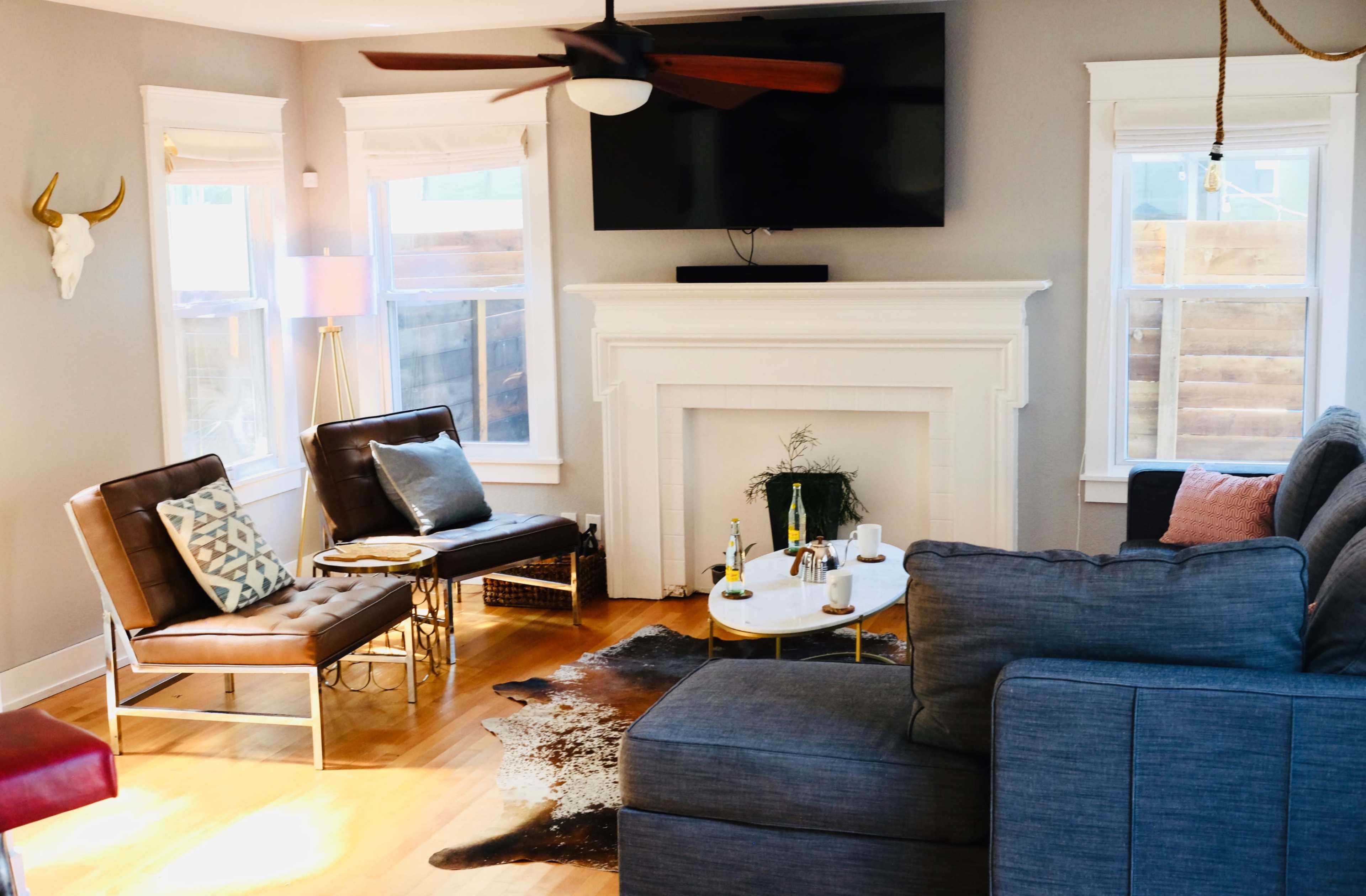 The image shows a modern living room featuring a sectional sofa, a round coffee table on a cowhide rug, and a flat-screen TV mounted above a white fireplace, with various seating arrangements and decor elements.