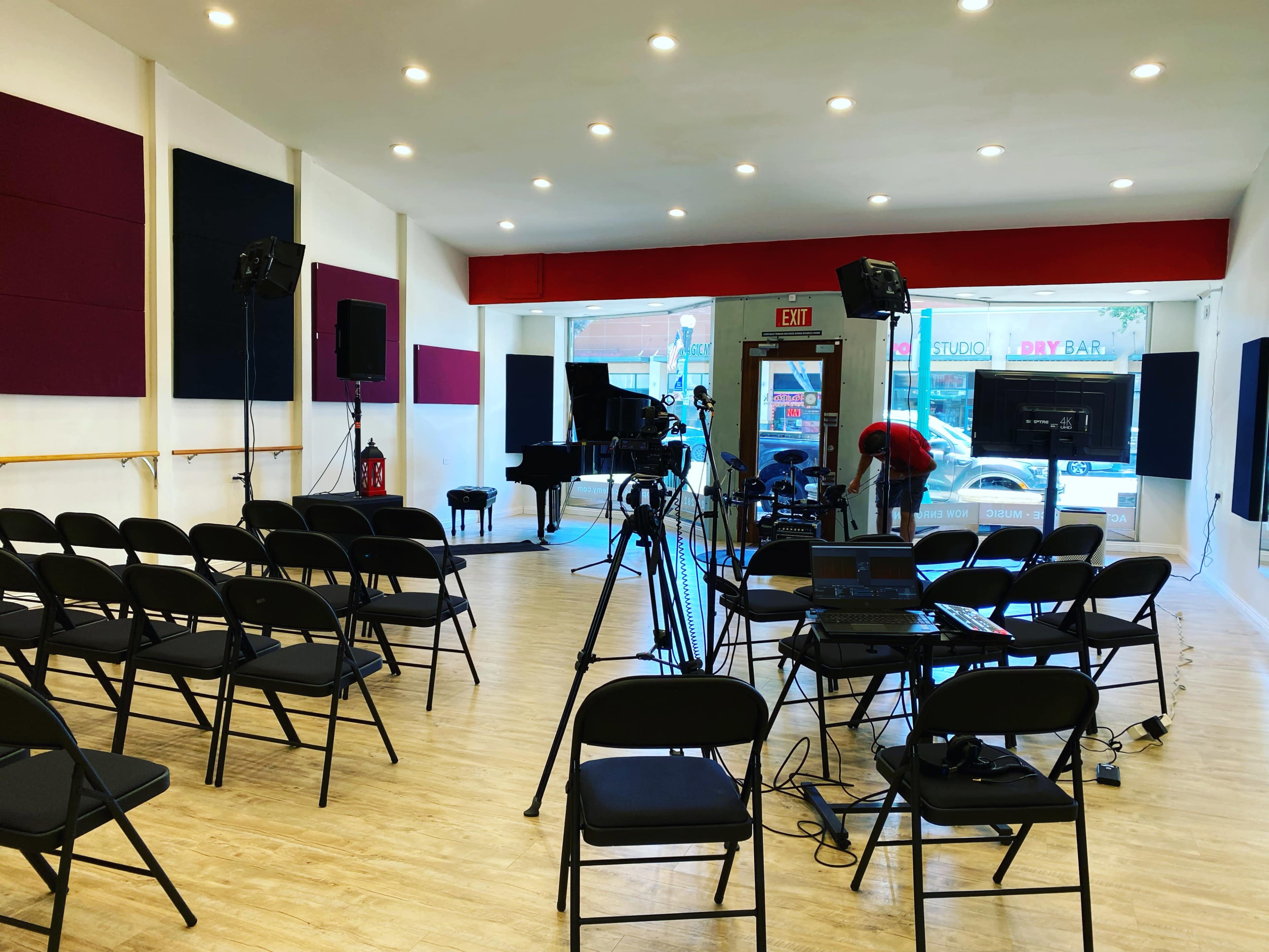 The scene shows a music studio setup with rows of black chairs facing a grand piano, surrounded by cameras and lighting equipment.