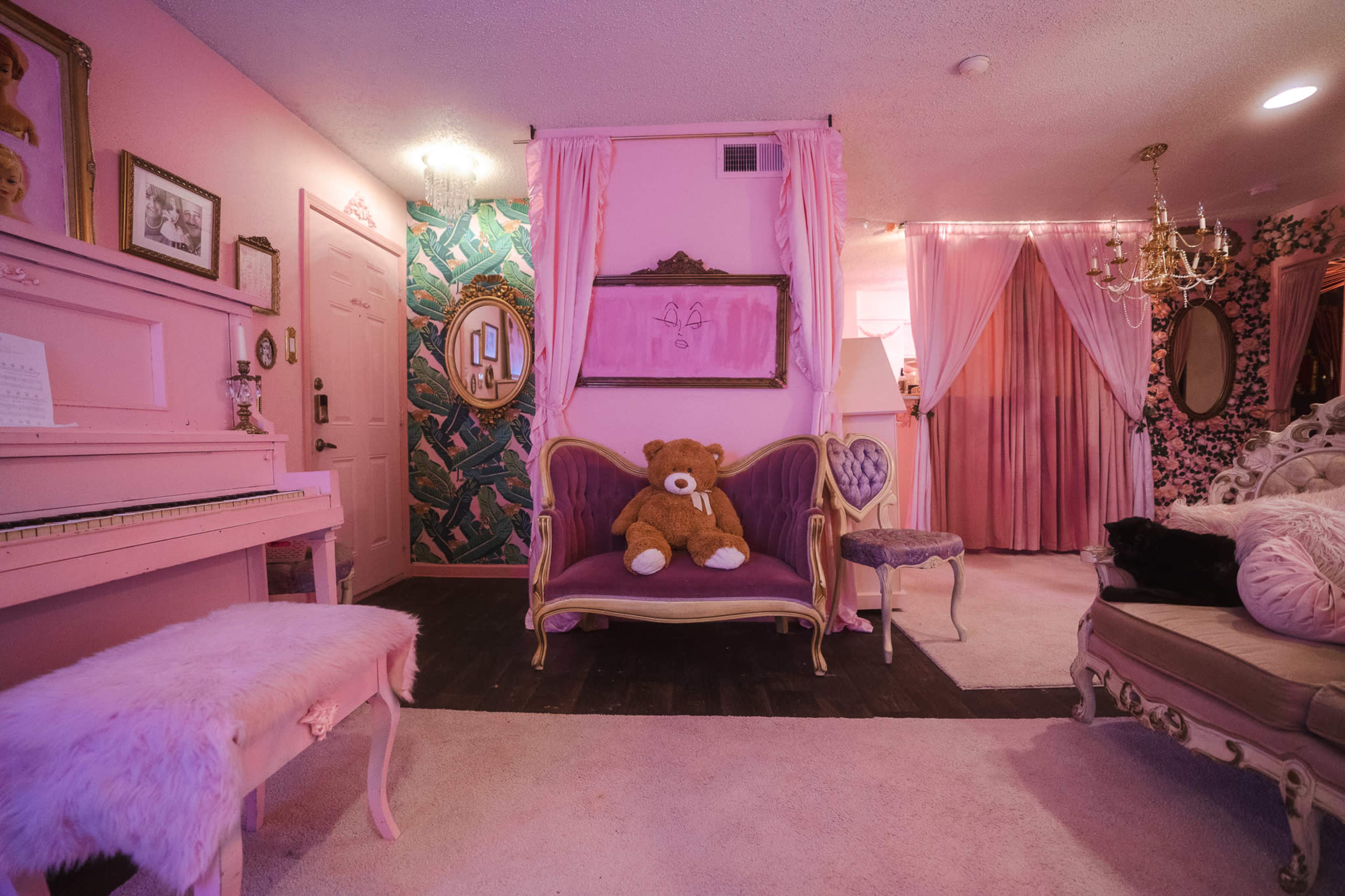 The room features pink walls, a vintage piano, a plush sofa with a teddy bear, and ornate furniture.