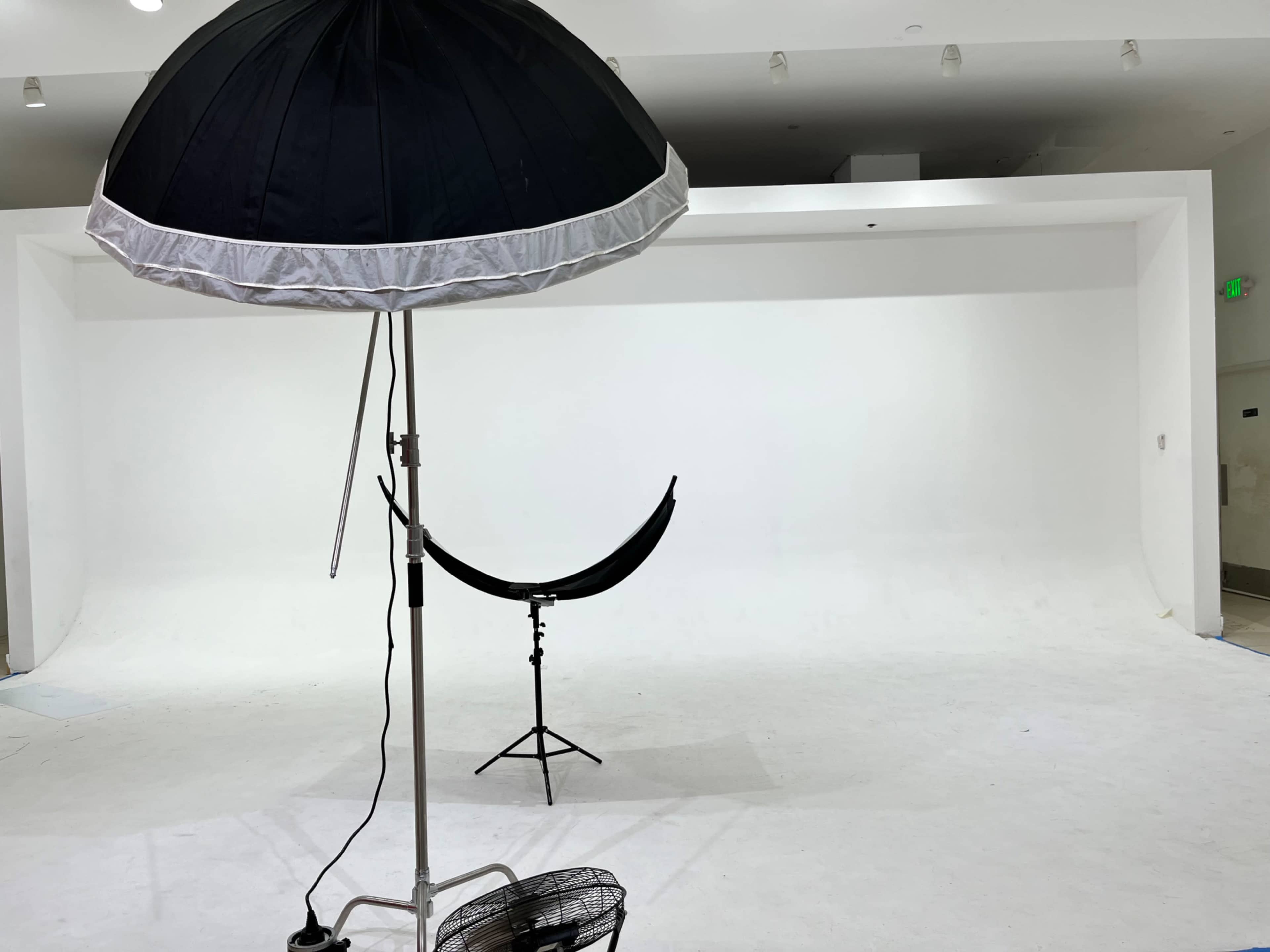The image shows a photography studio with a white backdrop, an umbrella light, and a fan.