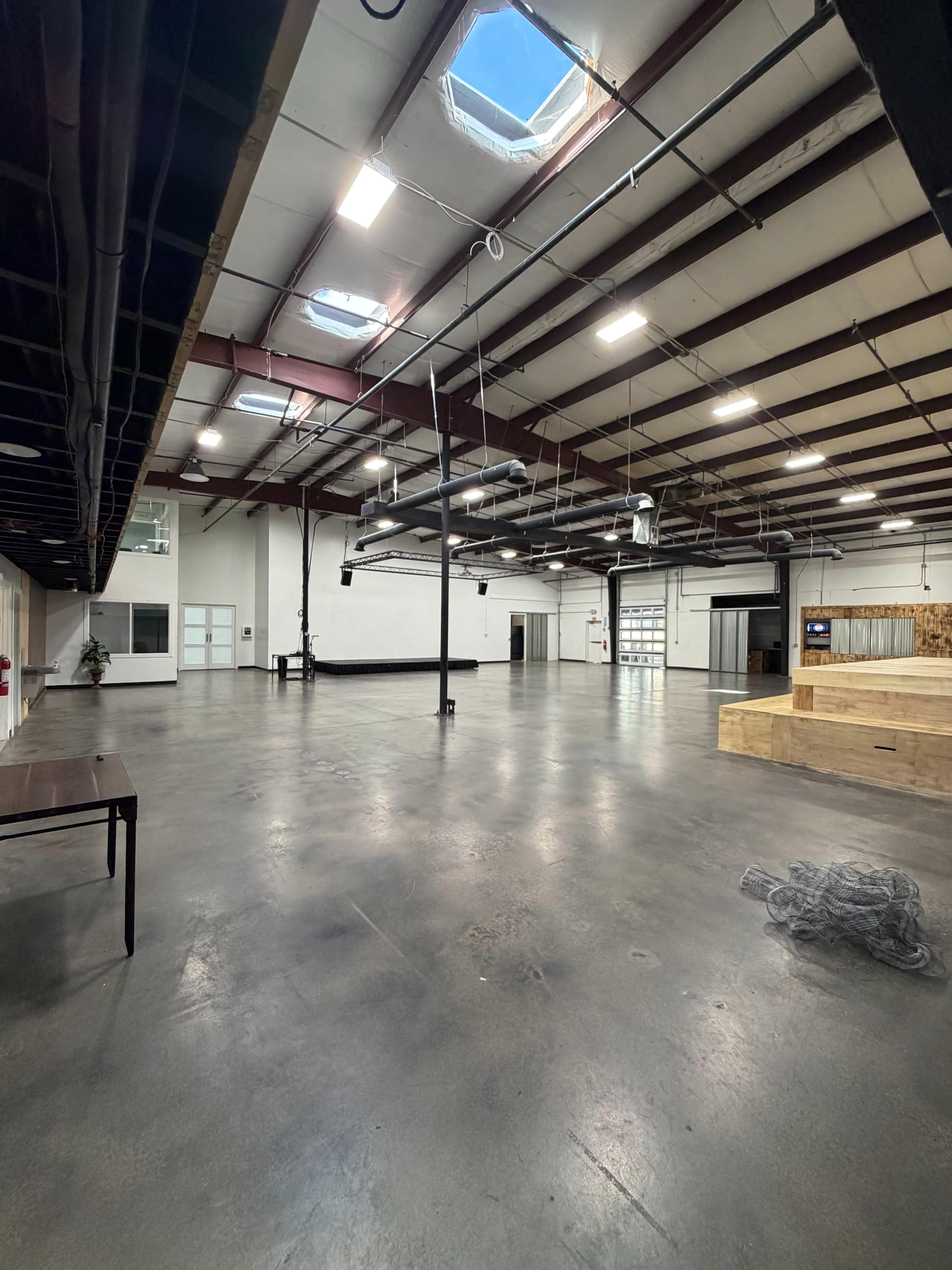 The image shows a spacious industrial interior with high ceilings, concrete floors, and various fixtures overhead, including skylights and metal beams.