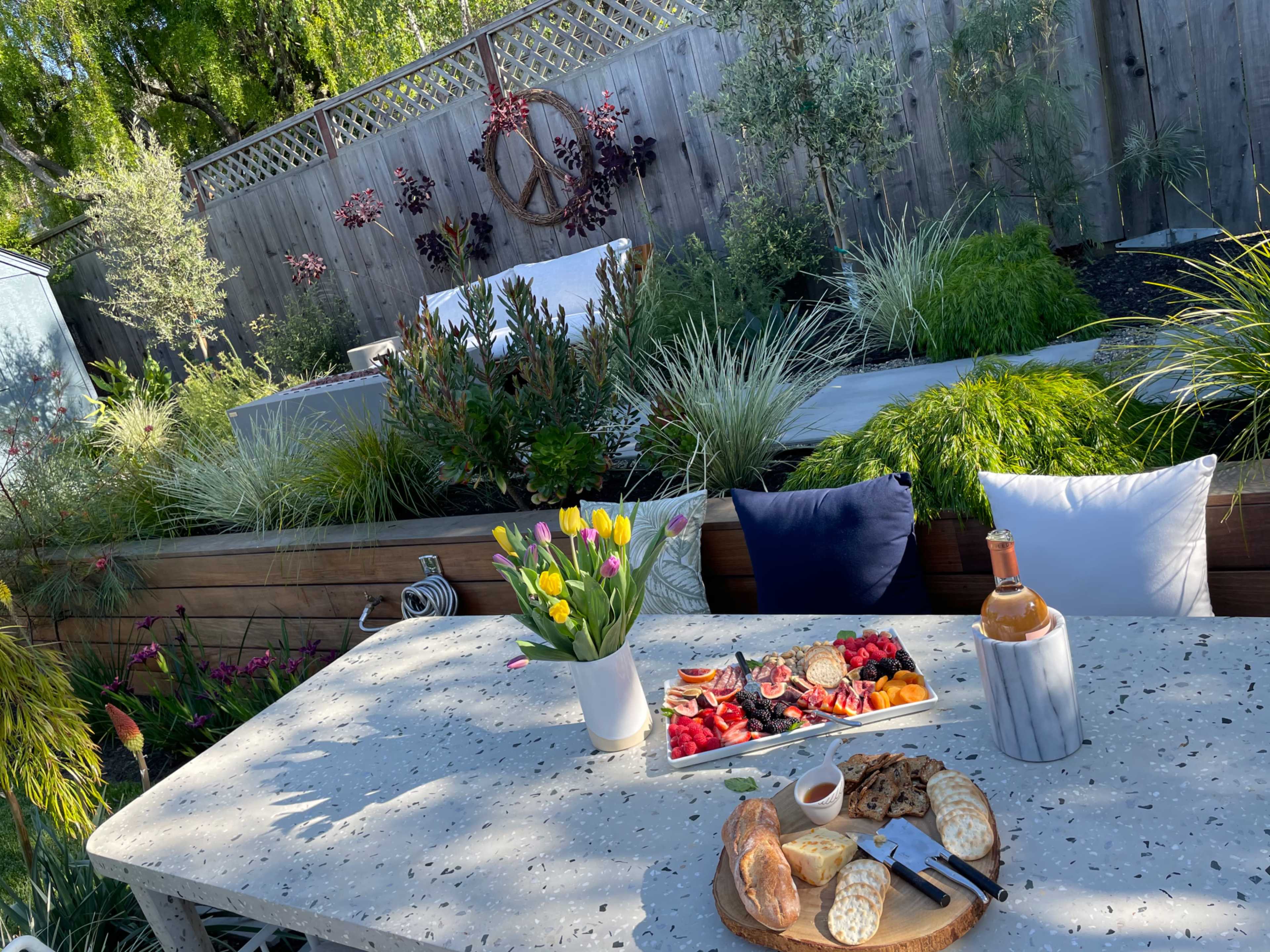 A stone table holds a platter of fruits, cheeses, and baked goods, surrounded by lush greenery and colorful flowers in a backyard setting.