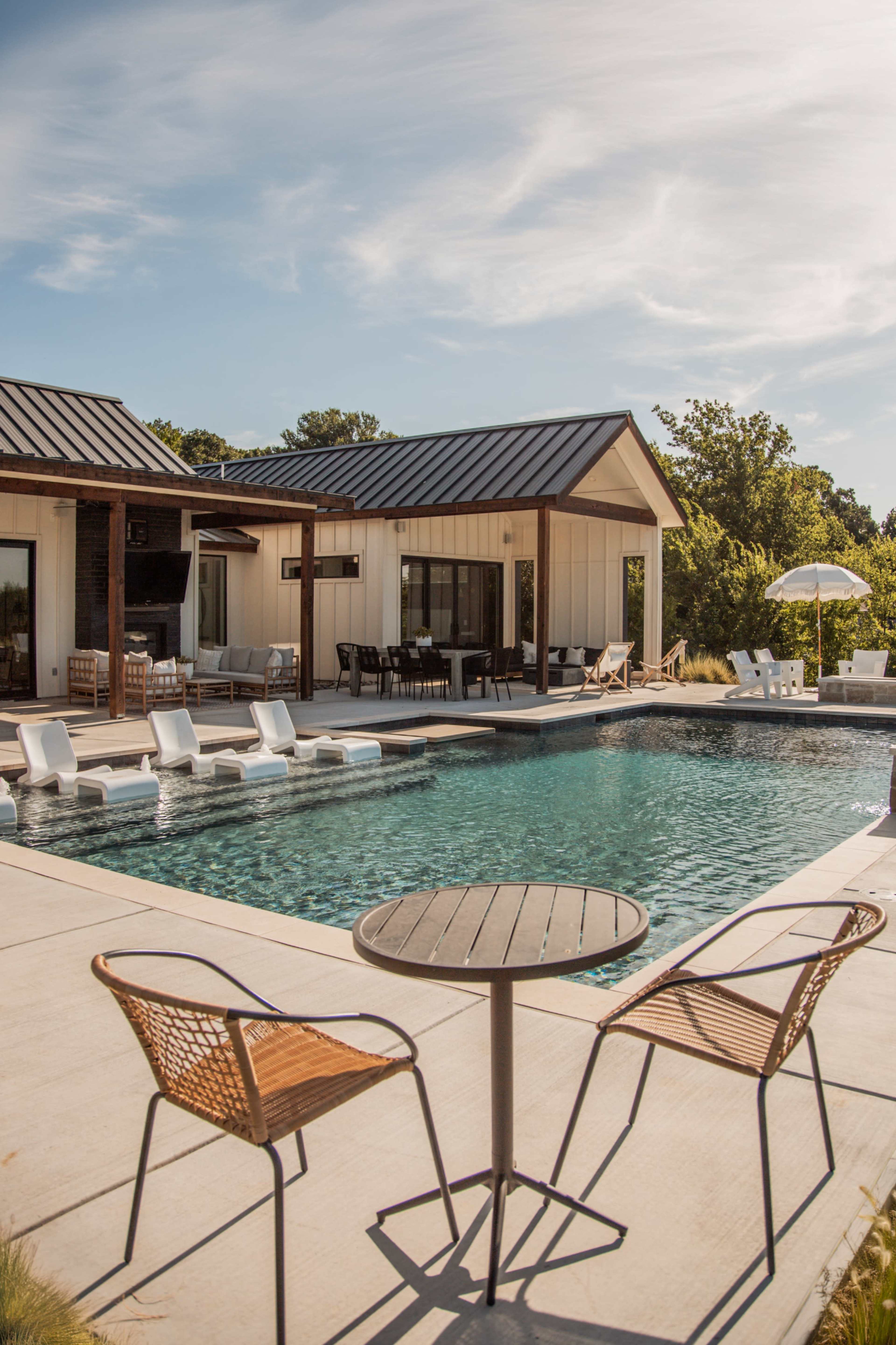 A modern backyard features a swimming pool with lounge chairs and a table, adjacent to a stylish house with a deck.
