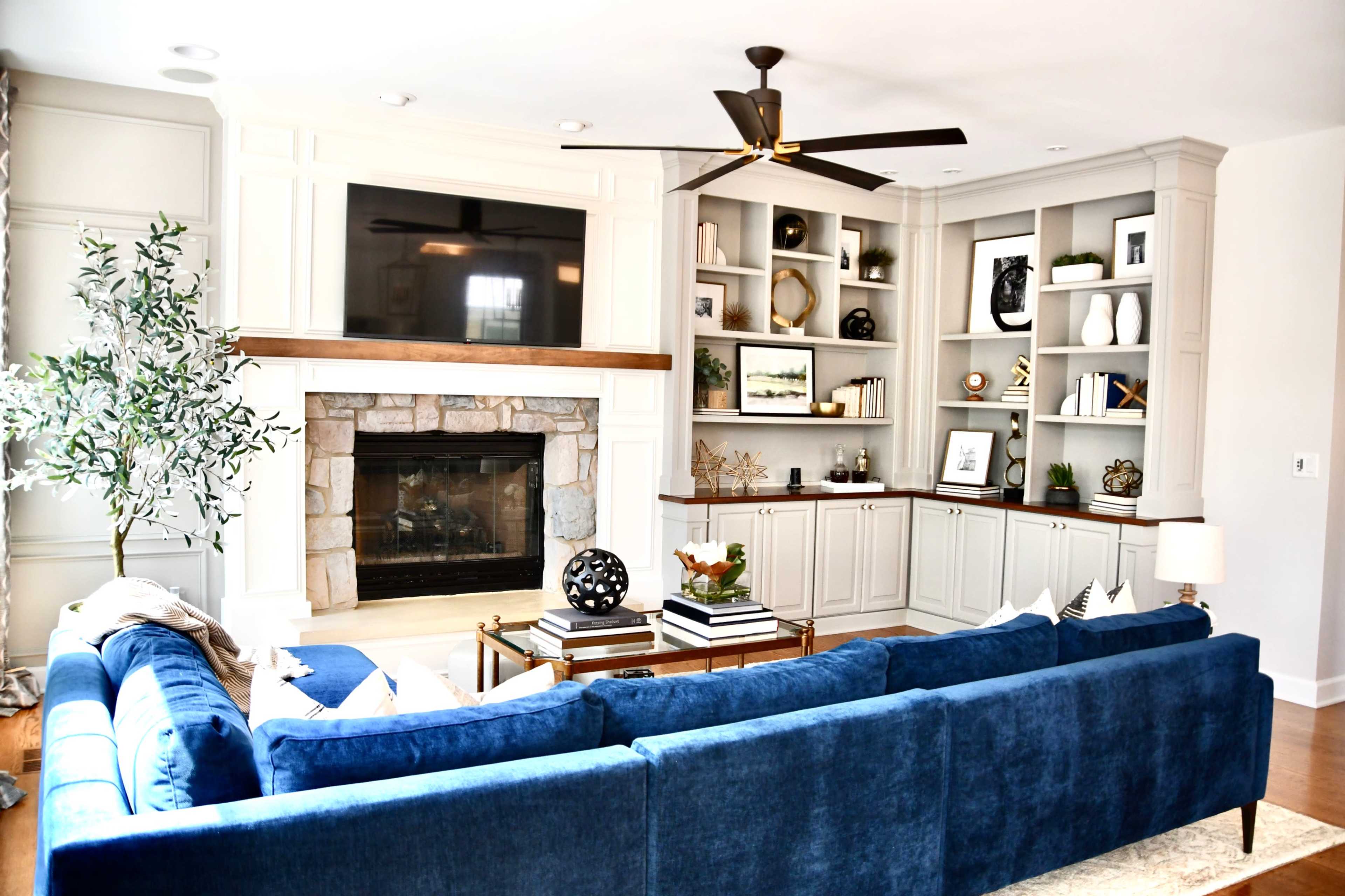 A spacious living room features a blue sectional sofa, a stone fireplace, built-in shelving with decorative items, and a ceiling fan.