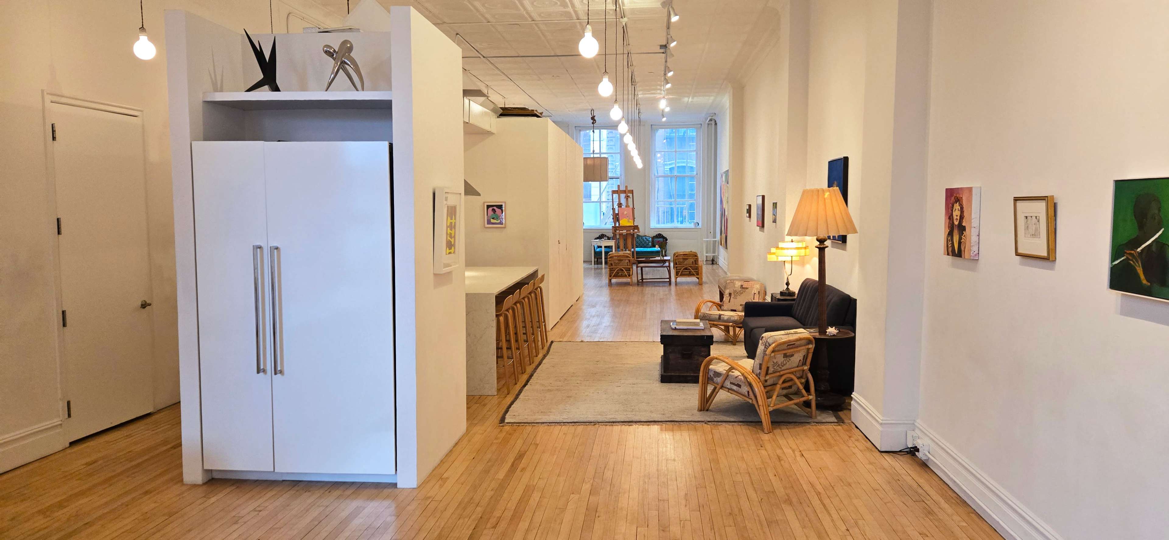 The image shows a spacious, modern apartment interior featuring an open layout with a kitchen area, a living space with a lamp and seating, and artwork displayed on the walls.