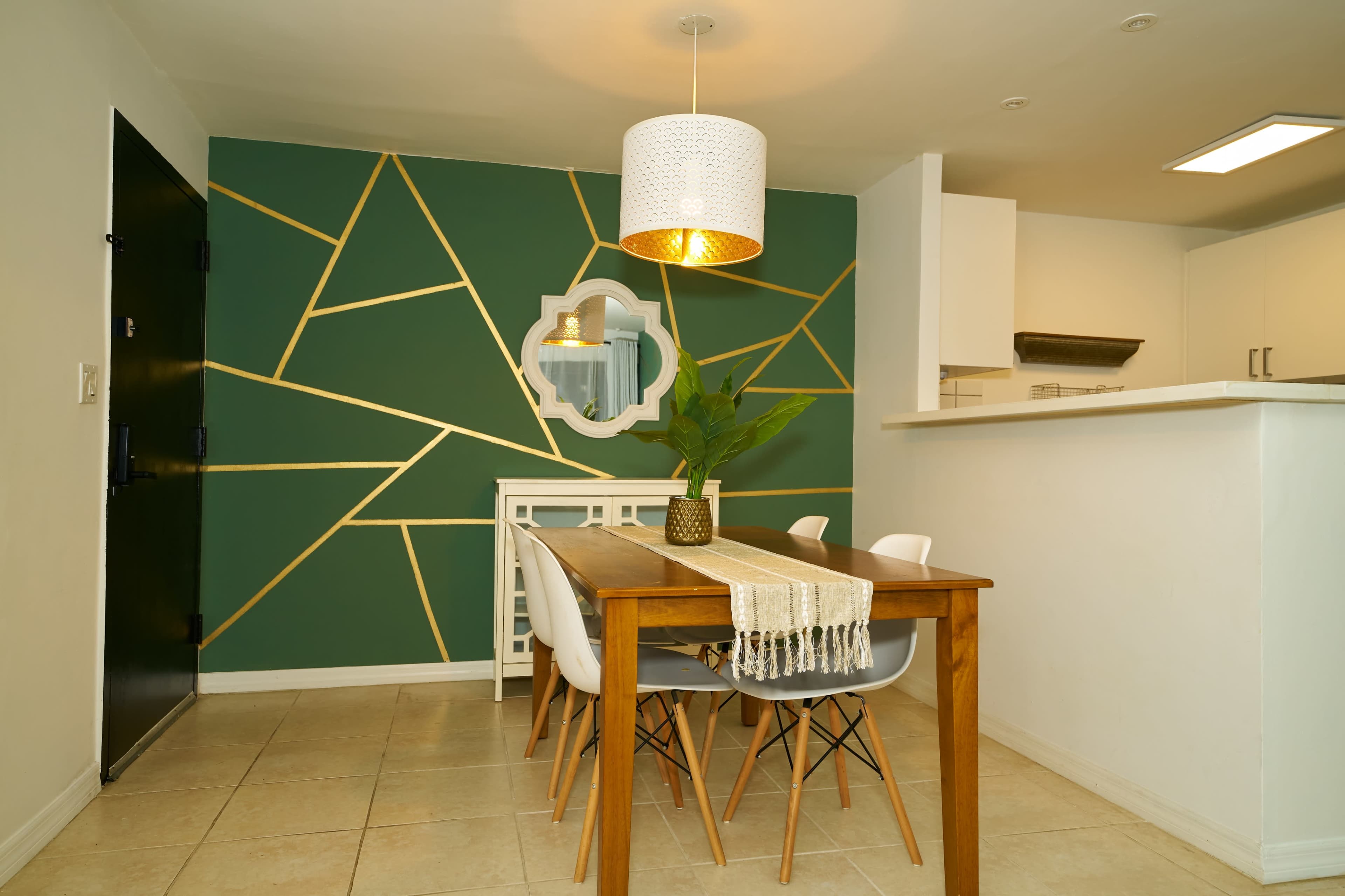 The image shows a modern dining area featuring a wooden table with white chairs, a decorative green wall with gold geometric lines, and a pendant light hanging above.