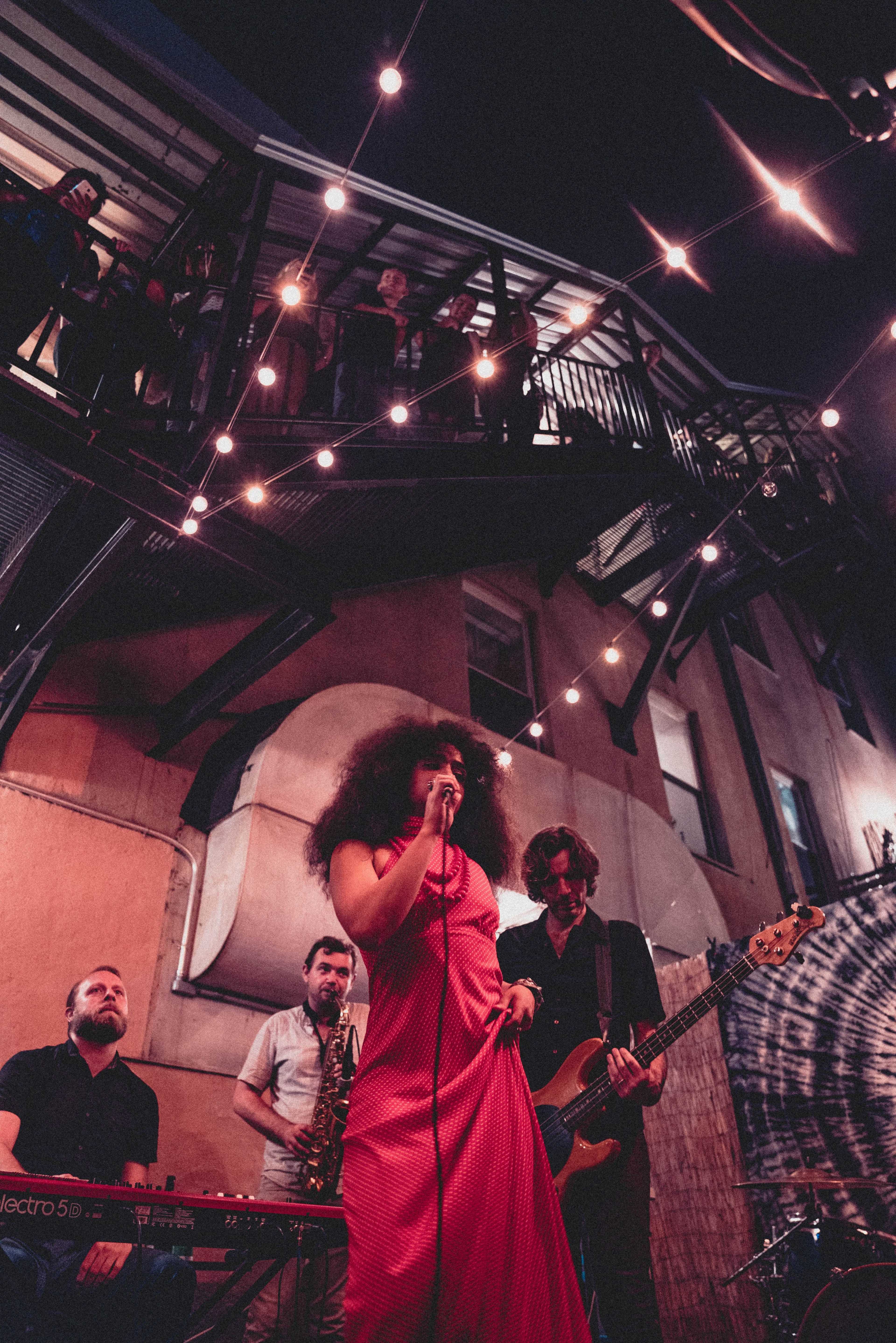 A woman in a red dress sings on stage while a band performs, and an audience watches from a balcony above.
