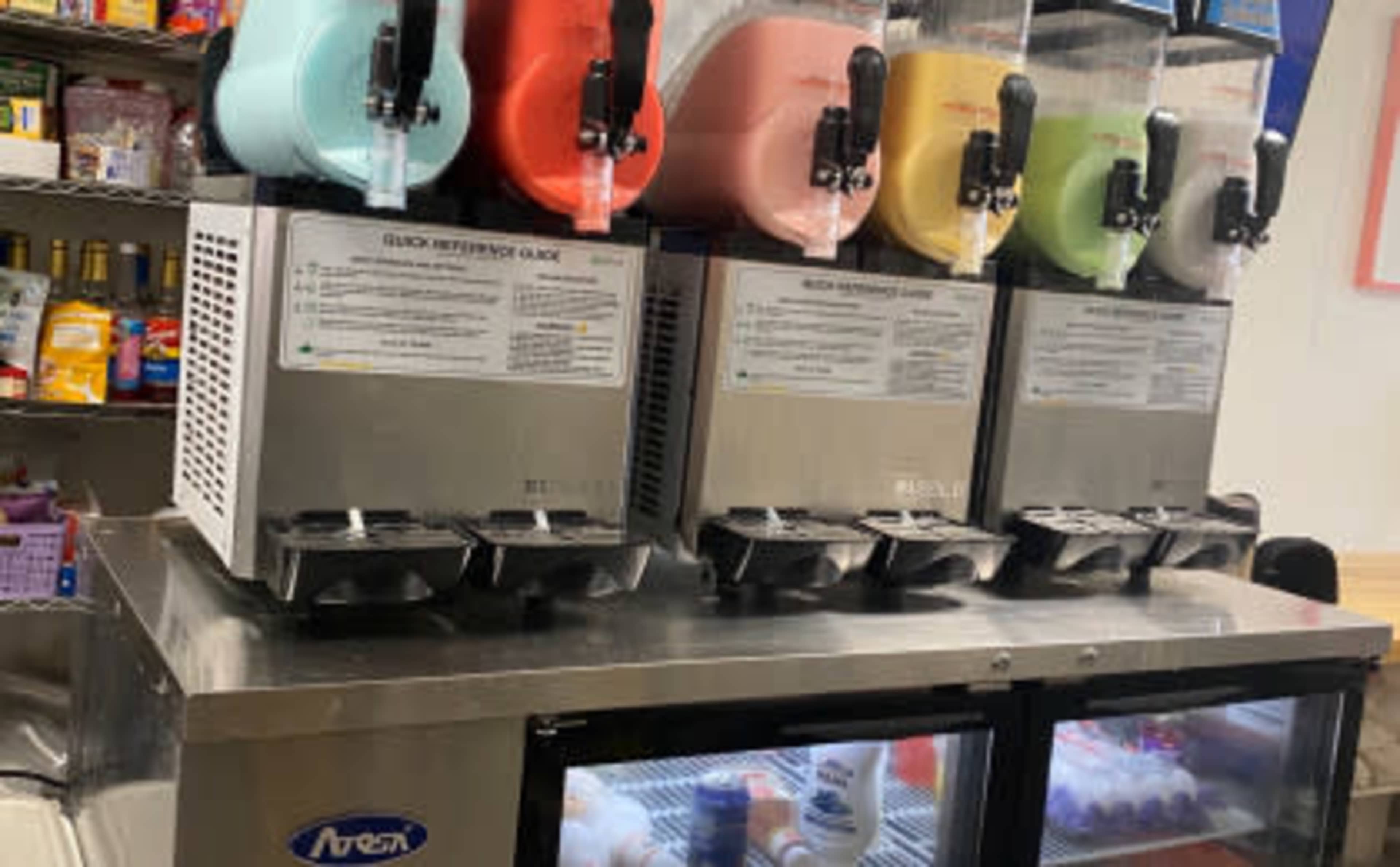 A countertop with three ice cream machines, each filled with different colored flavors, alongside a glass-front refrigerator.