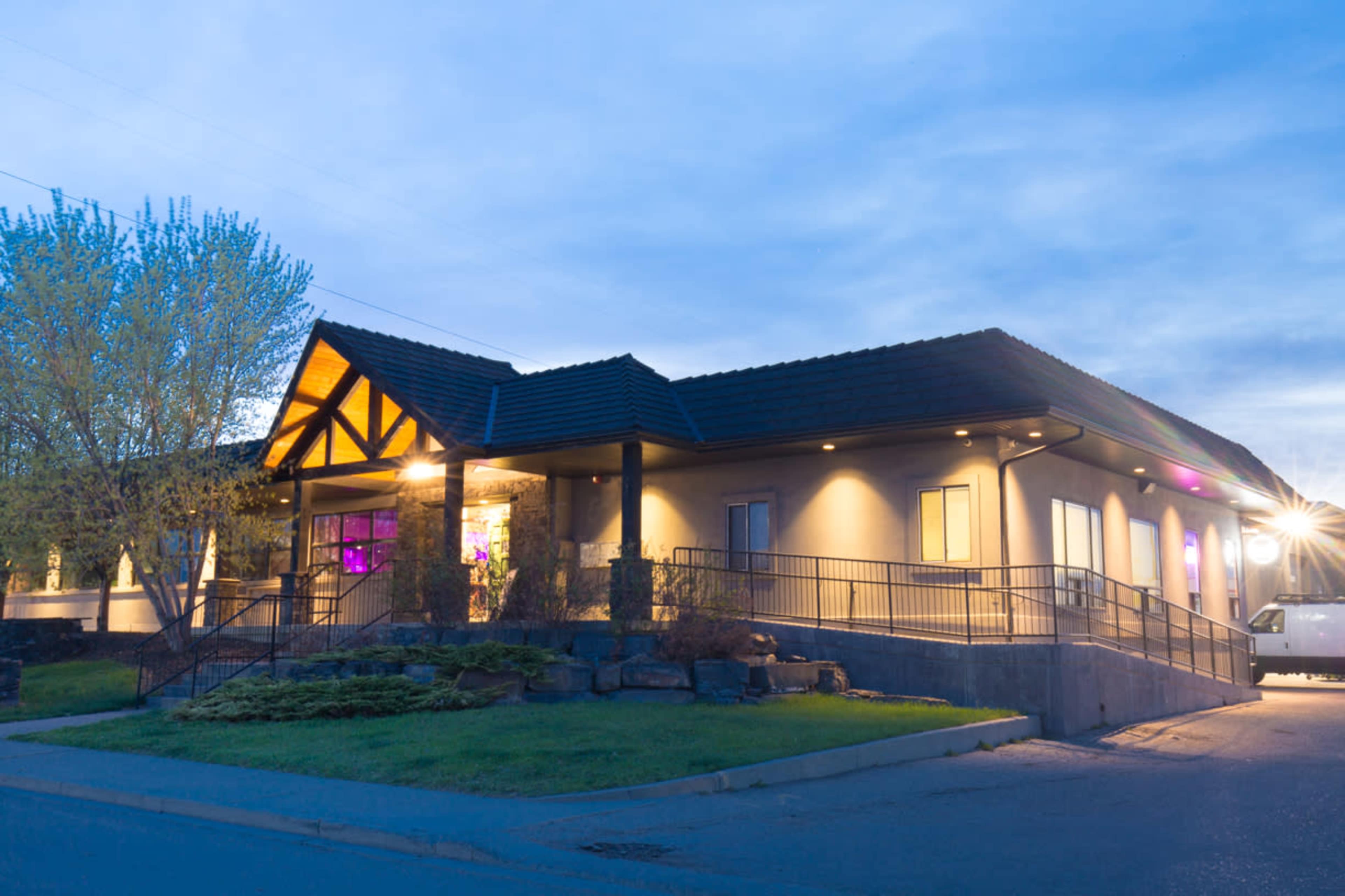 A modern building with a sloped roof, a ramp leading to the entrance, and illuminated windows under a twilight sky.