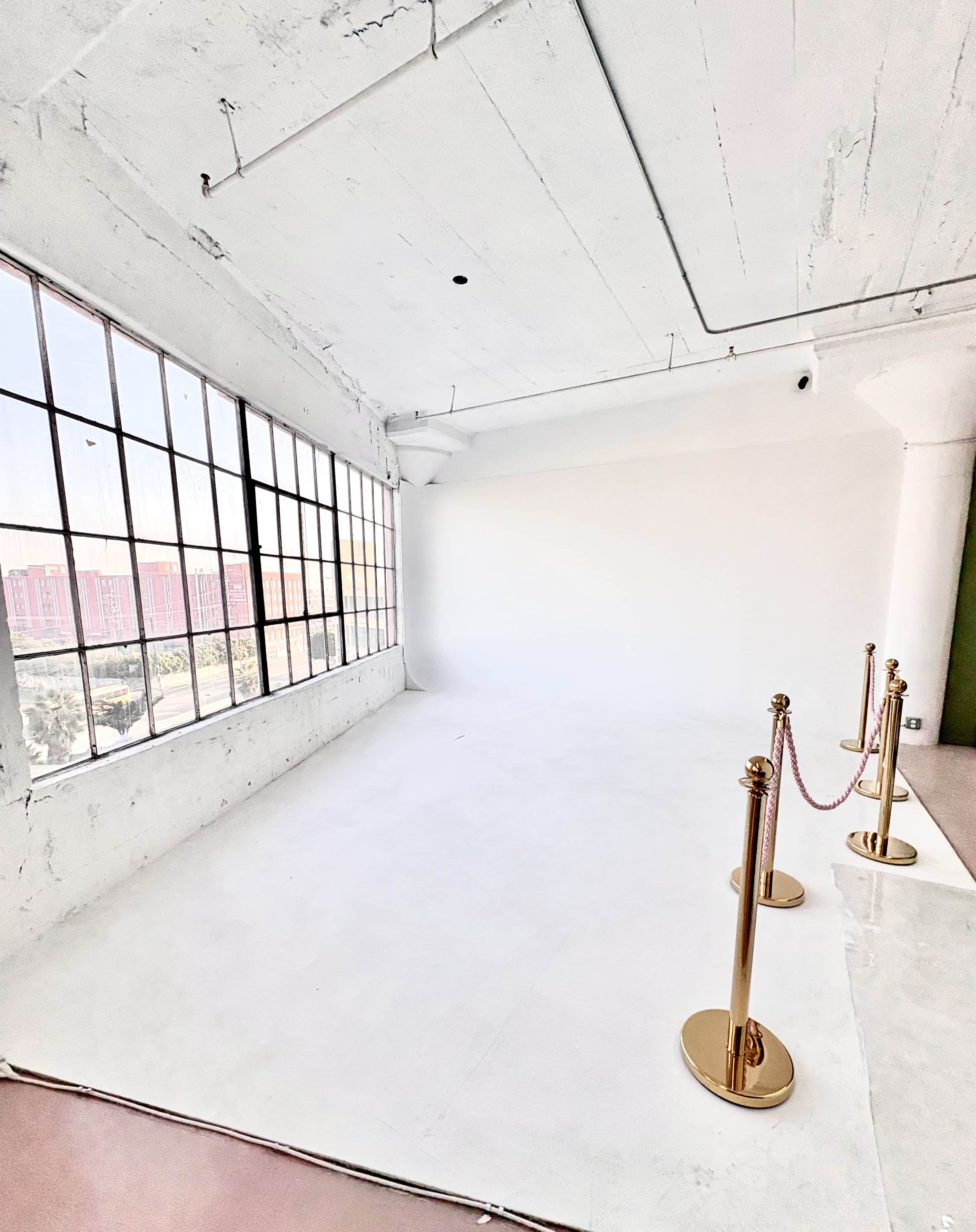 The image shows a spacious, empty room with a large window allowing natural light to enter, and two gold stanchions with a red rope positioned on the floor.