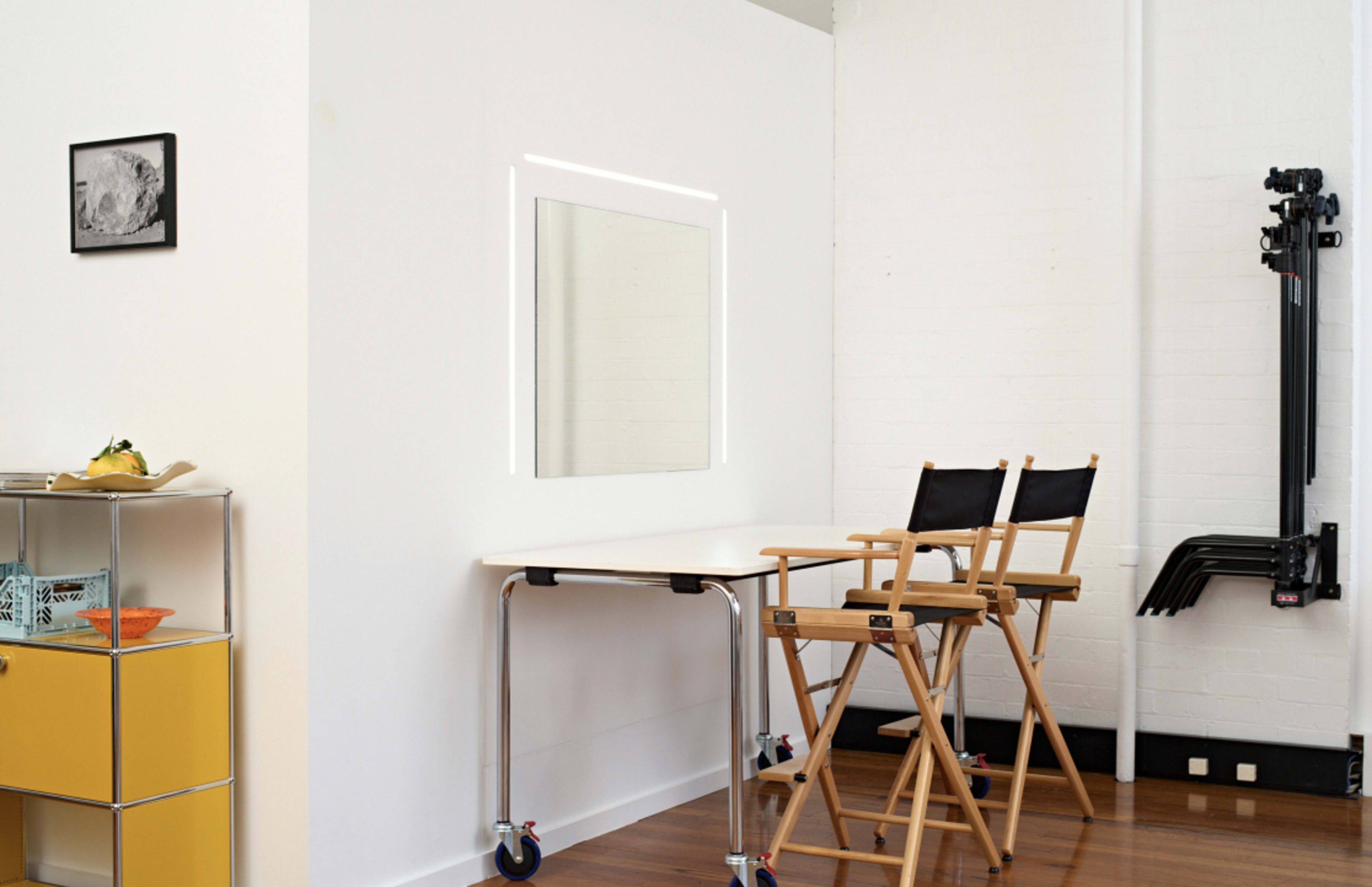 A minimalist room with a wall-mounted mirror, two director's chairs, a folding table, and a wall-mounted rack holding equipment.