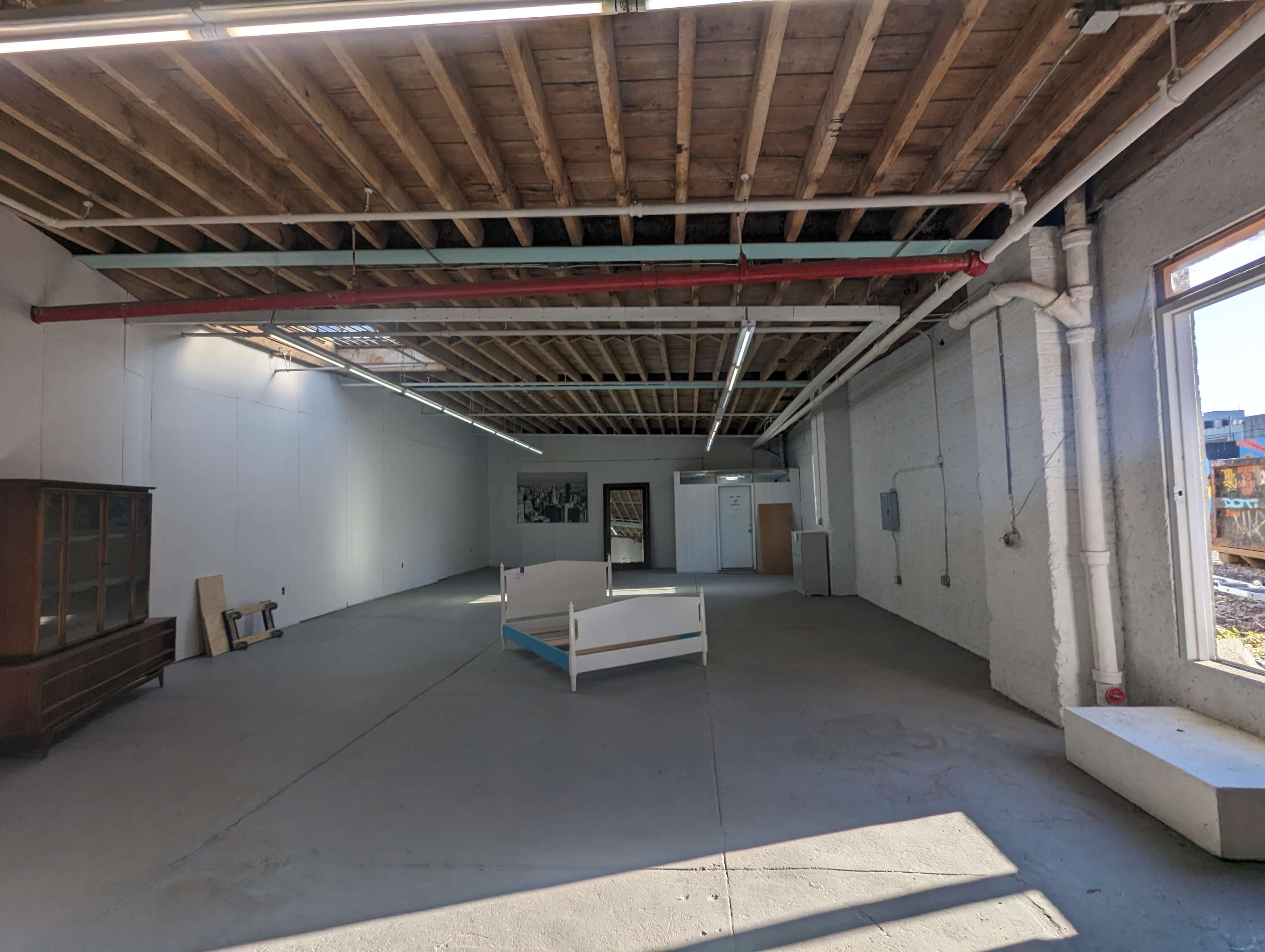 The image shows a spacious, empty room with exposed wooden beams and concrete flooring, featuring a bed frame in the center and a large window on one side.