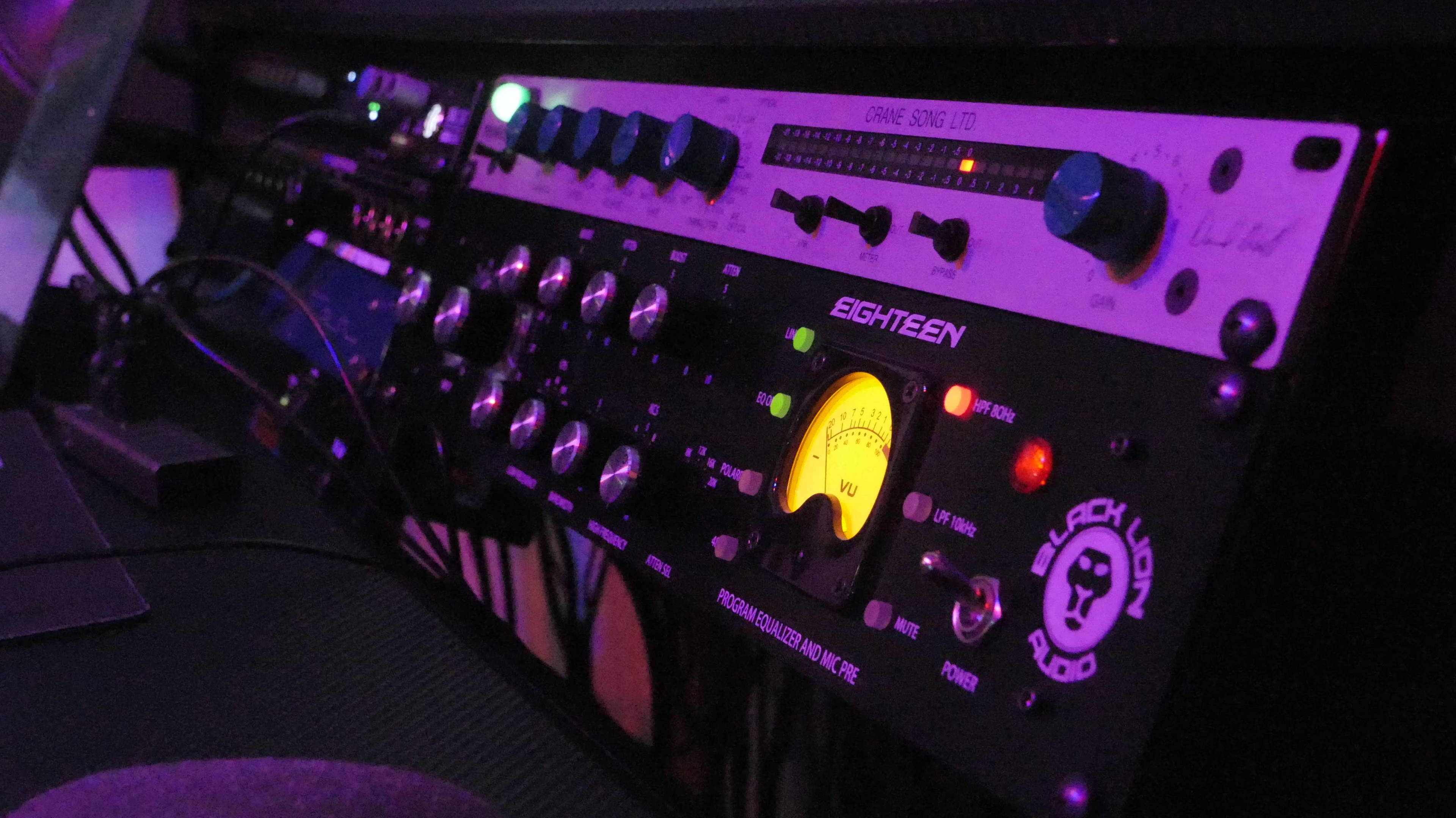 The image shows a close-up of audio recording equipment, featuring various knobs, buttons, and a VU meter displaying signal levels.