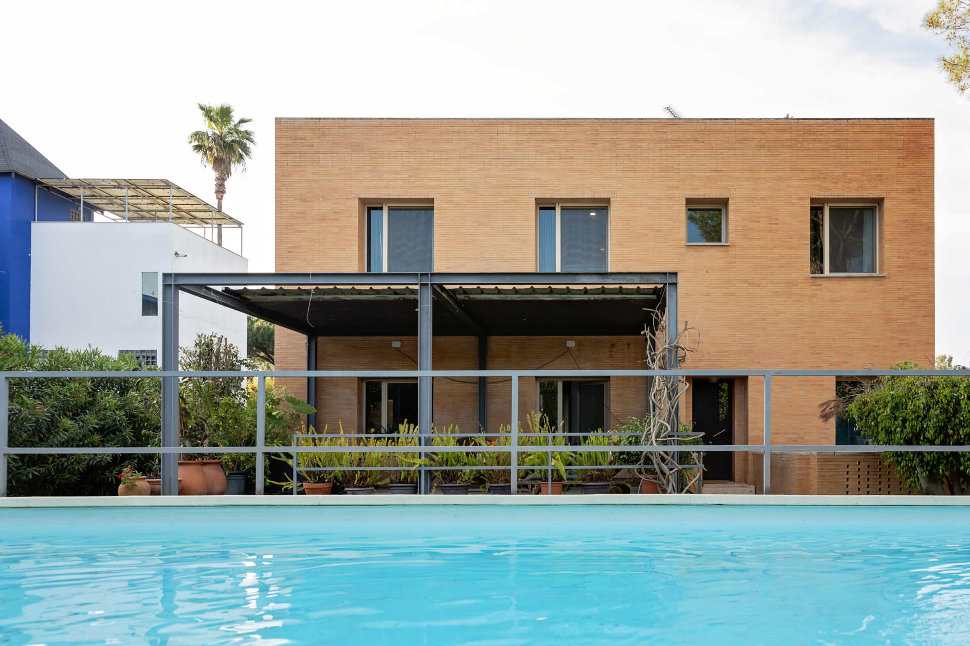 The image showcases a modern two-story building with a brick exterior, featuring a shaded patio and a swimming pool in the foreground.