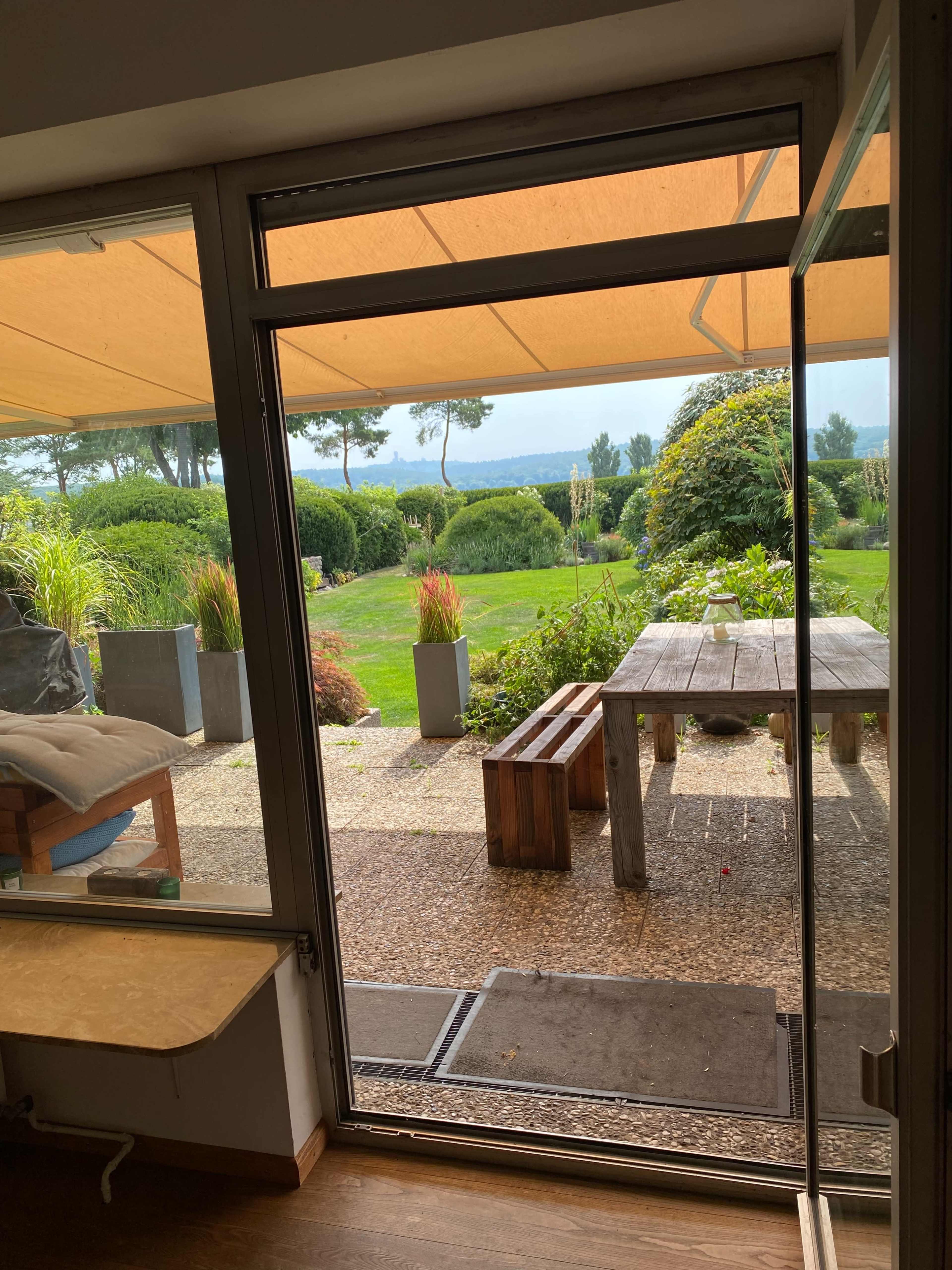 A view from inside a room through a glass door, leading to a garden with a wooden table, benches, and landscaped greenery.