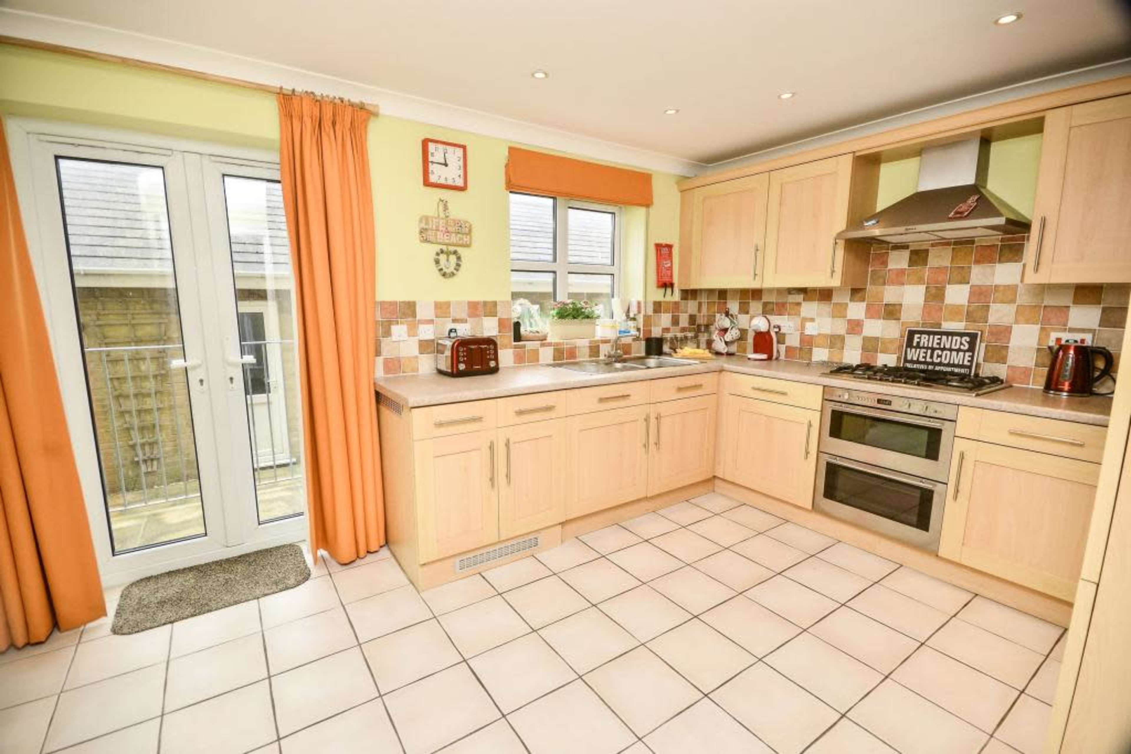 A modern kitchen with light wooden cabinetry, tiled flooring, and large windows allowing natural light.