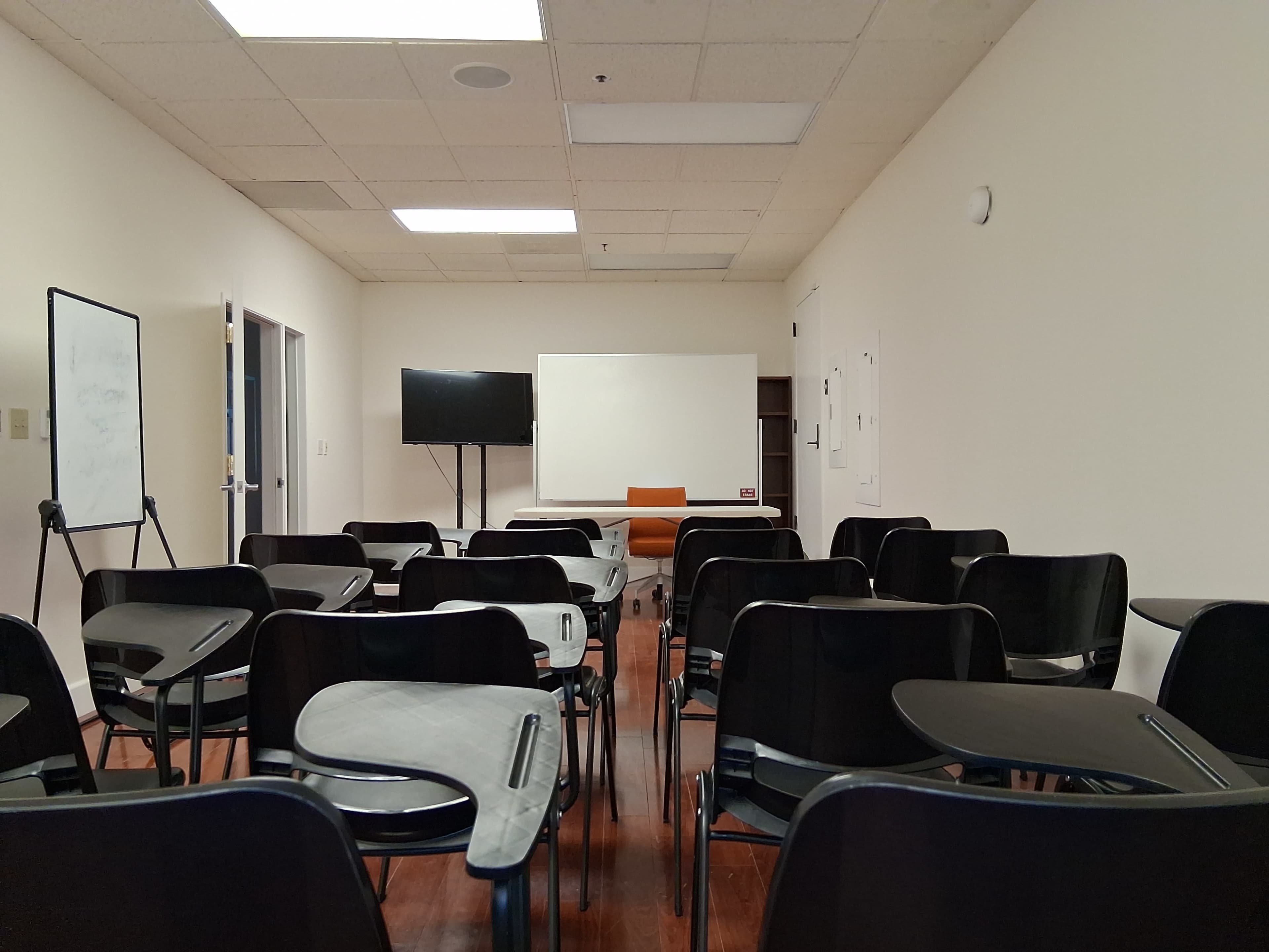 The image shows an empty classroom with rows of black desks facing a presentation screen and an orange chair at the front.