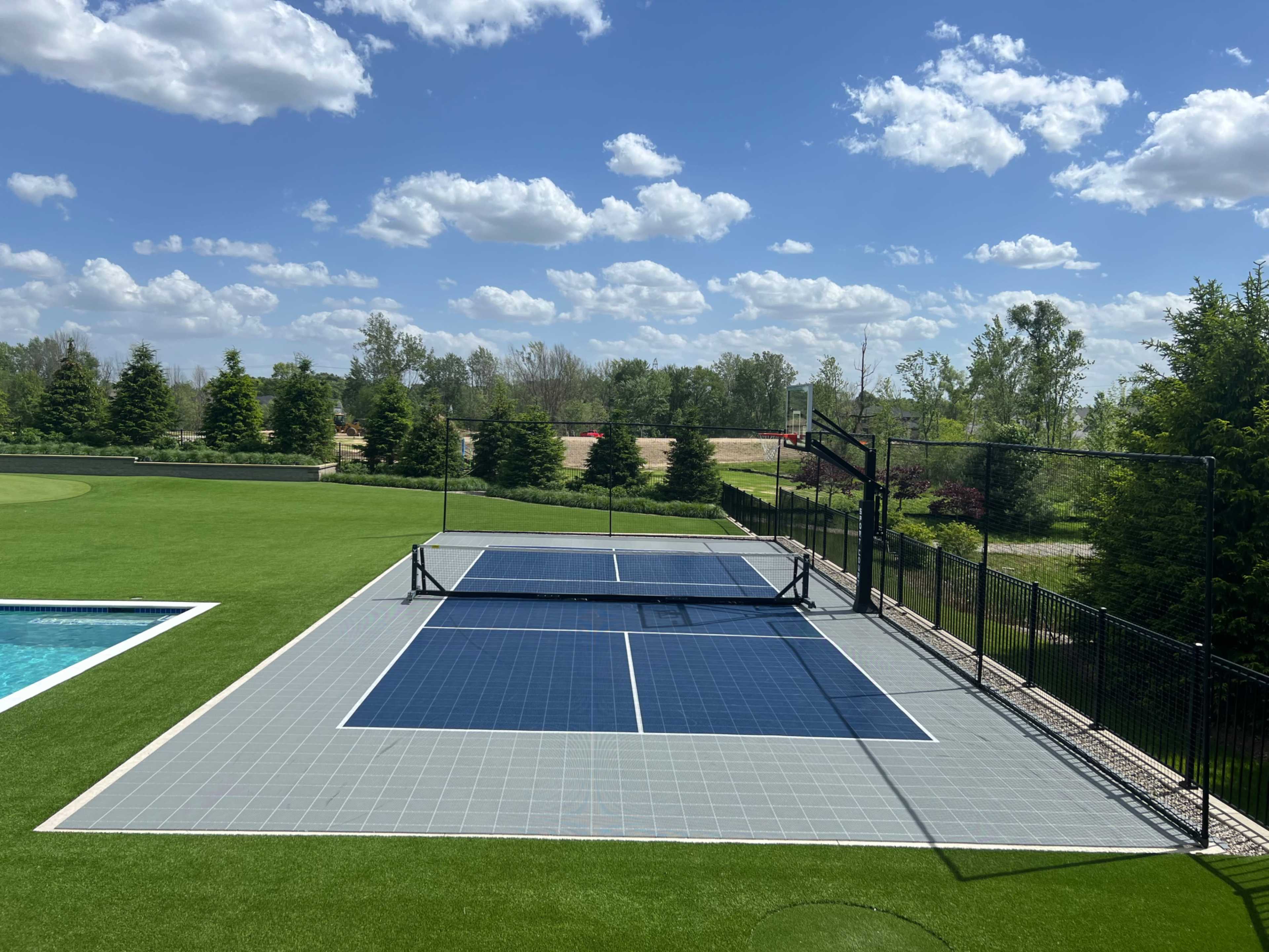 A tennis court with solar panels sits next to a swimming pool, surrounded by grass and trees under a partly cloudy sky.