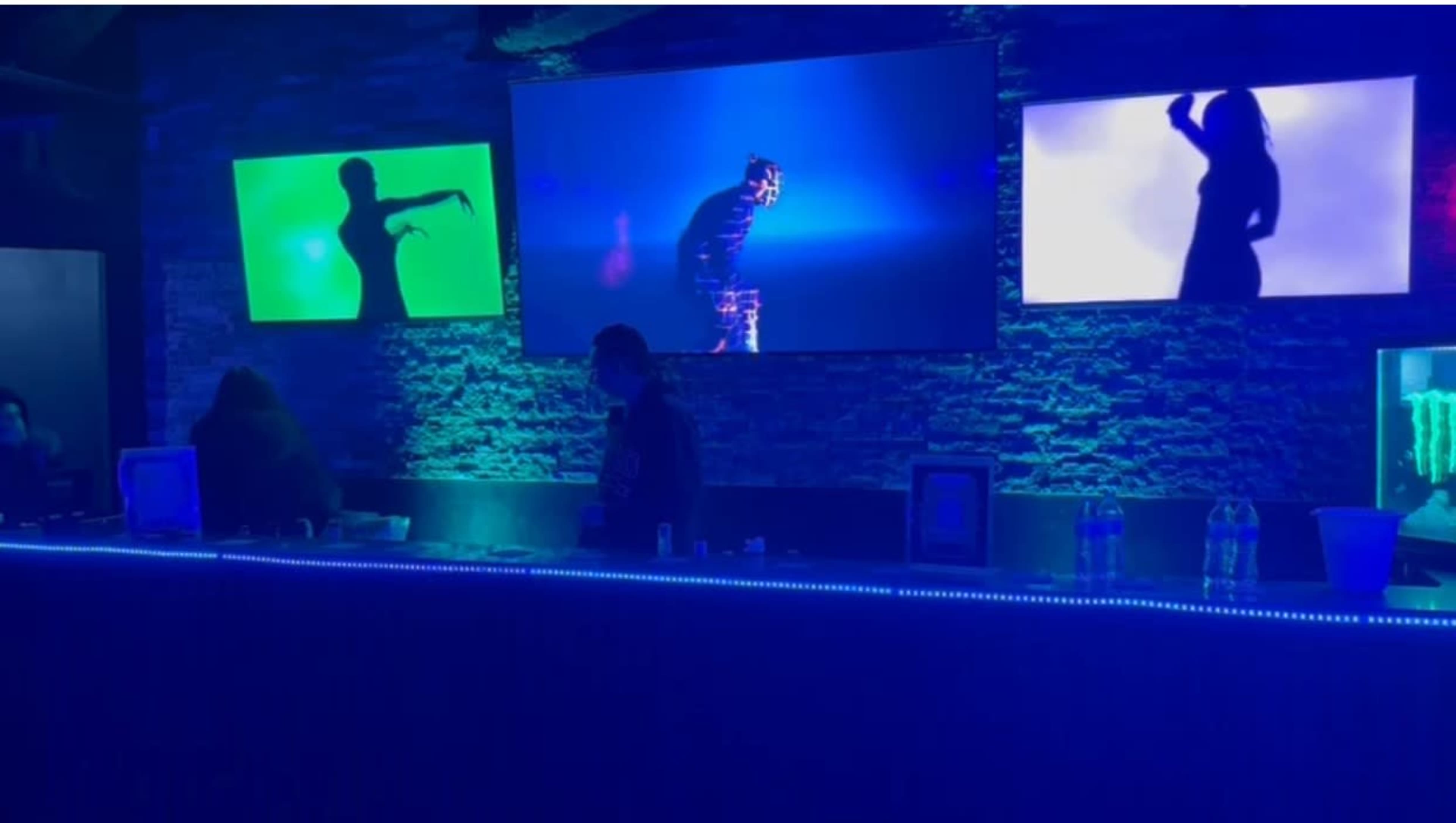 The image shows a bar area with illuminated screens displaying silhouettes against a backdrop of blue and green lighting.