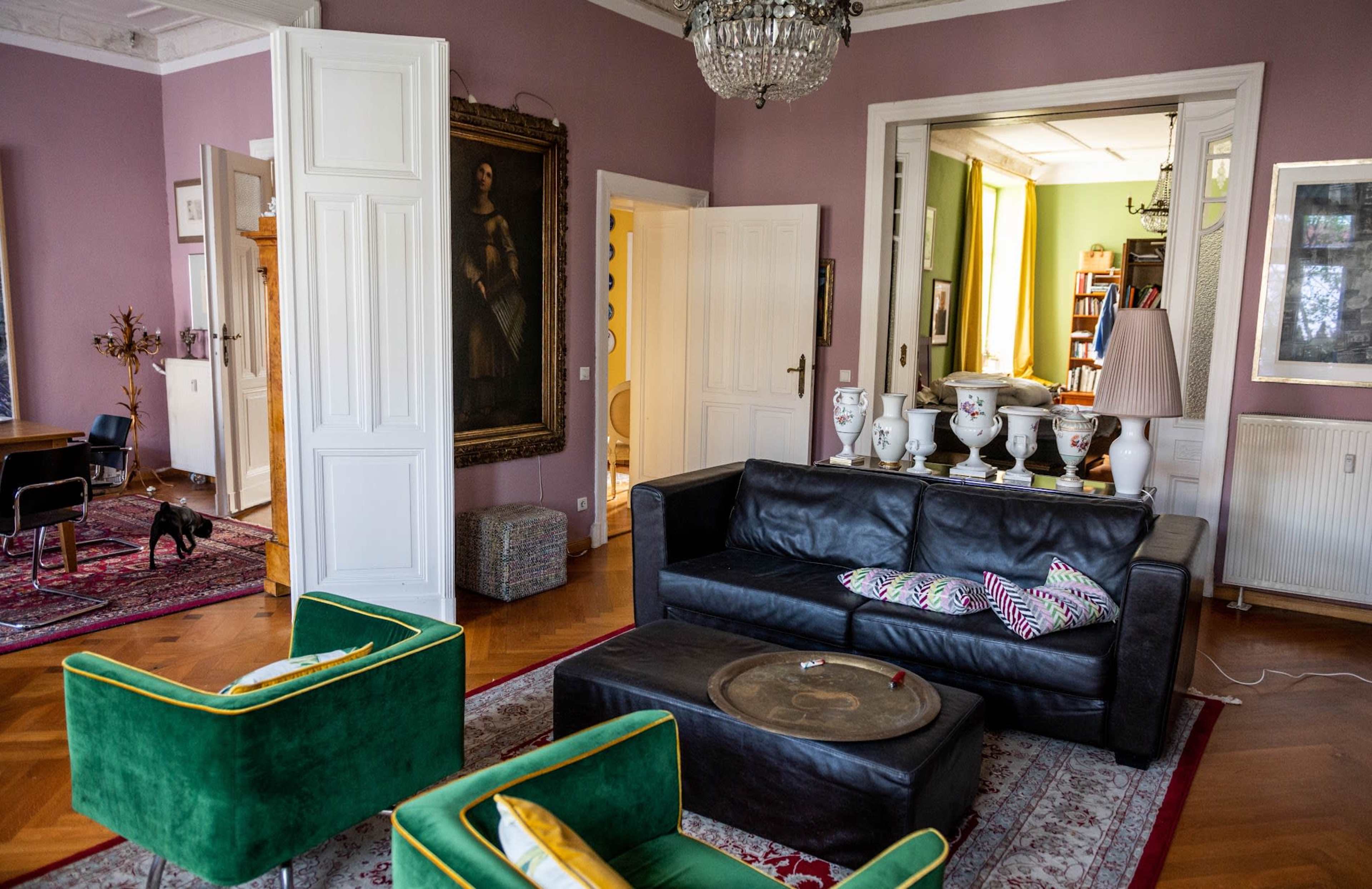 The image shows a stylish living room with a black leather sofa, two green armchairs, a wooden table, and various decorative items, including a chandelier and a large painting on the wall.