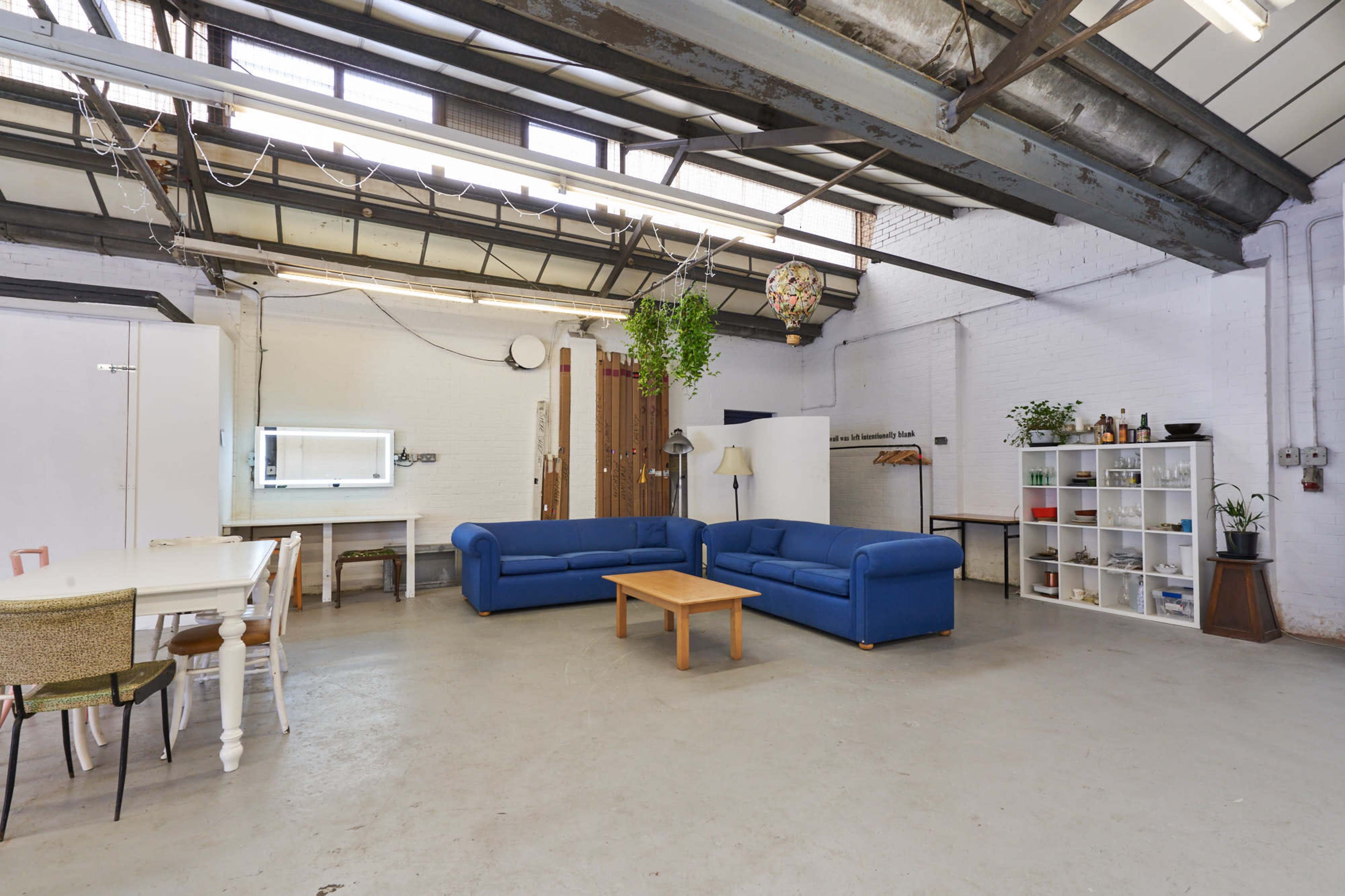 A spacious industrial-style room features two blue sofas, a wooden coffee table, and a dining area with chairs, alongside shelves and a large window.