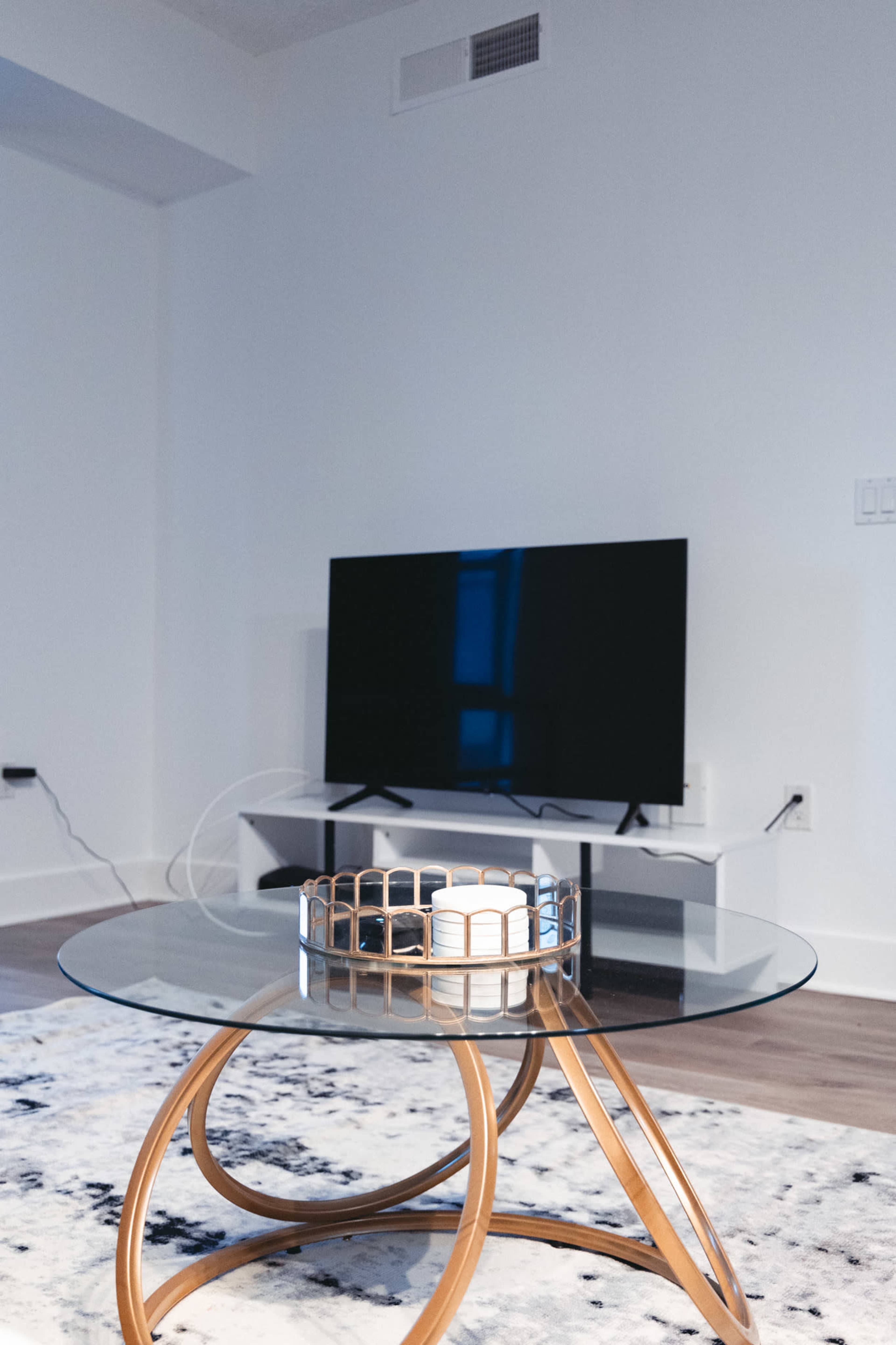 A modern living room features a glass-top coffee table with a decorative tray and a wall-mounted television in the background.